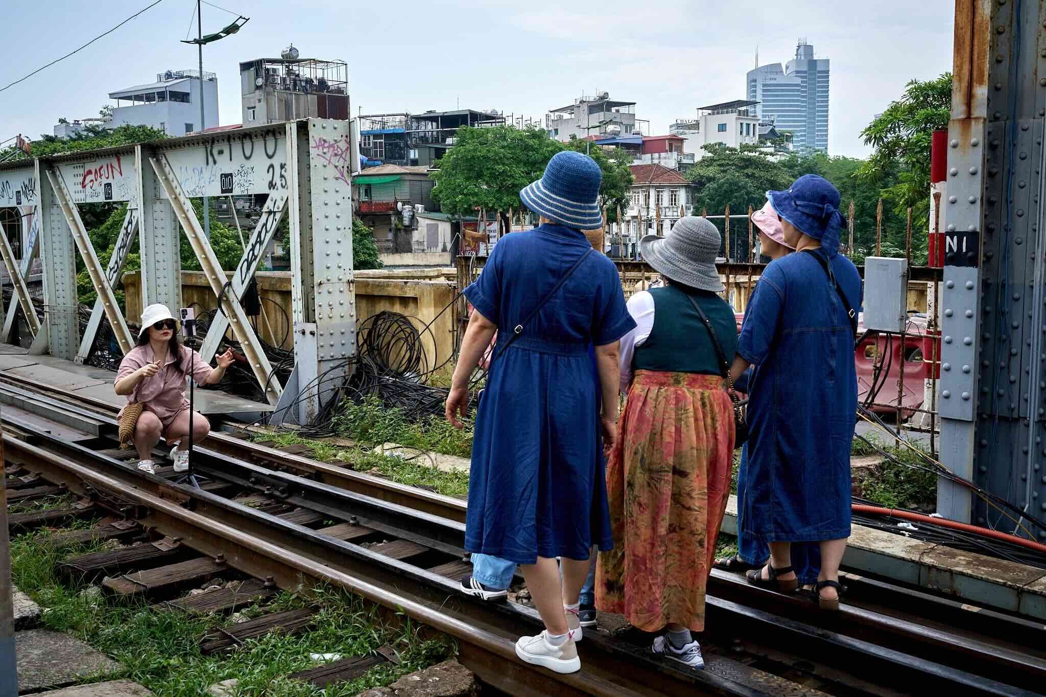 Group of tourists standing on a railway track, wearing blue and pink hats, observing a cityscape with residential buildings and a high-rise tower in the background.