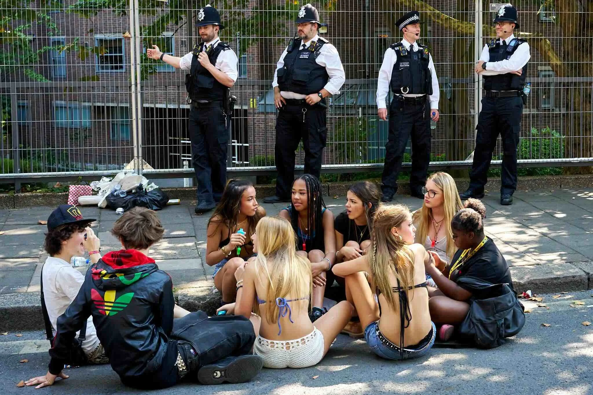 Group of young people sitting on the ground in front of a fence, talking and socializing, with four police officers standing behind the fence watching.