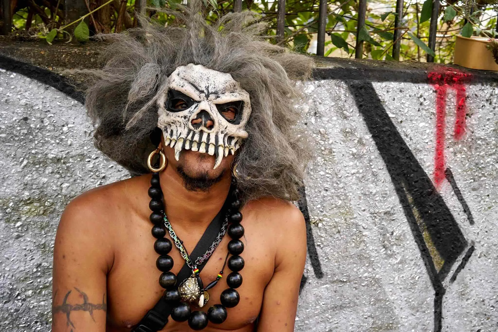 Person wearing a detailed skull mask with long, gray hair, large gold hoop earrings, and multiple black necklaces, standing outdoors next to a graffiti-covered wall with green foliage and a metal fence in the background.