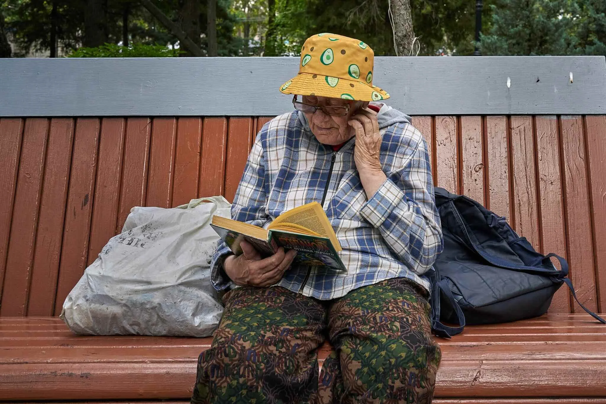 Jerzy Jaworek, Street and Travel Photography, Kazakhstan, Almaty. An elderly woman wearing a yellow hat with avocado patterns, glasses, and a plaid shirt sits on a red wooden bench, reading a paperback book. She has a large white plastic bag and a ba