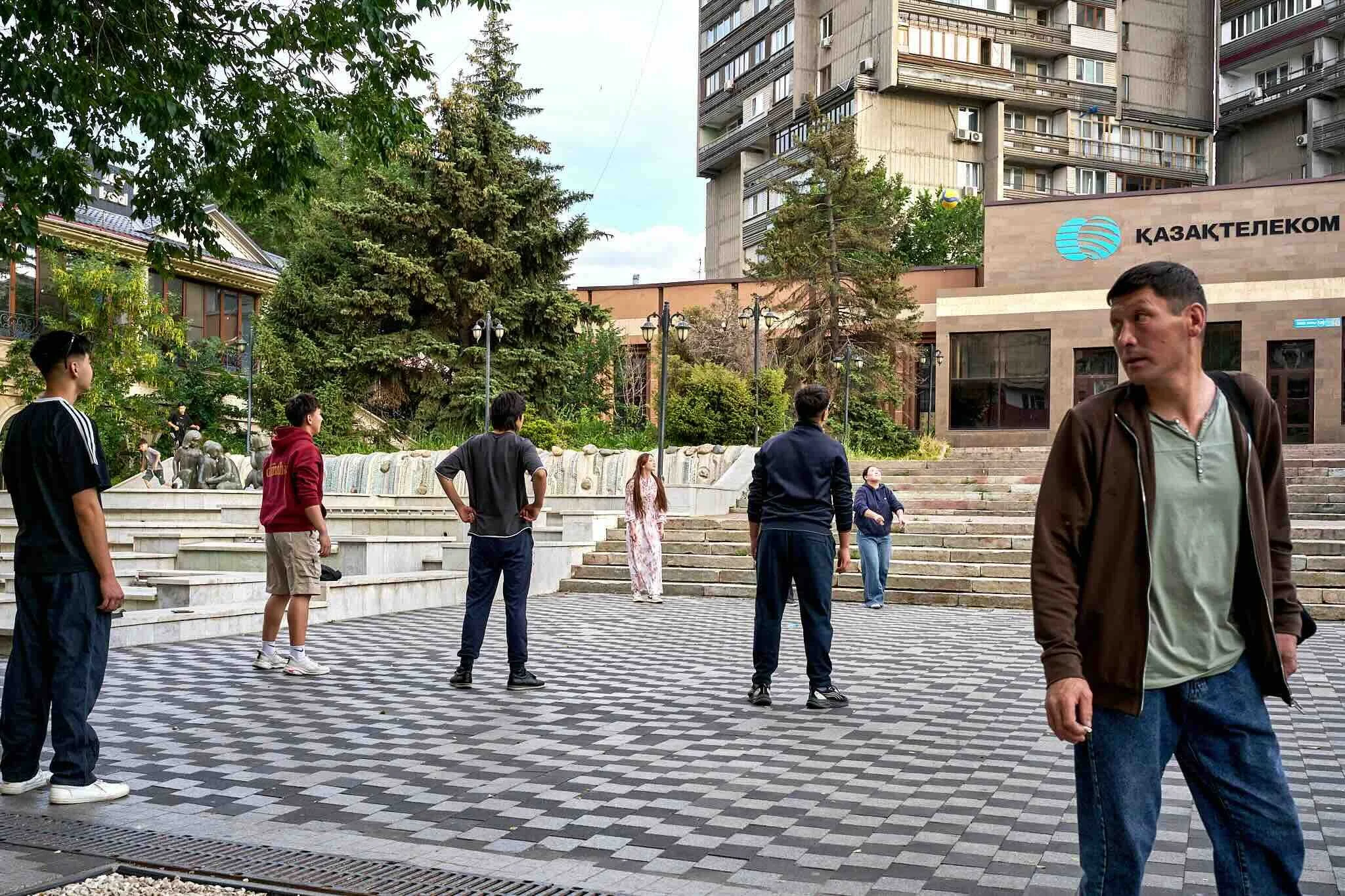 Jerzy Jaworek, Street and Travel Photography, Kazakhstan, Almaty. Group of people gathered in an urban park or plaza with trees, stairs, and a fountain, surrounded by tall buildings, with a sign that reads 'KAZAKTELECOM' on one building.