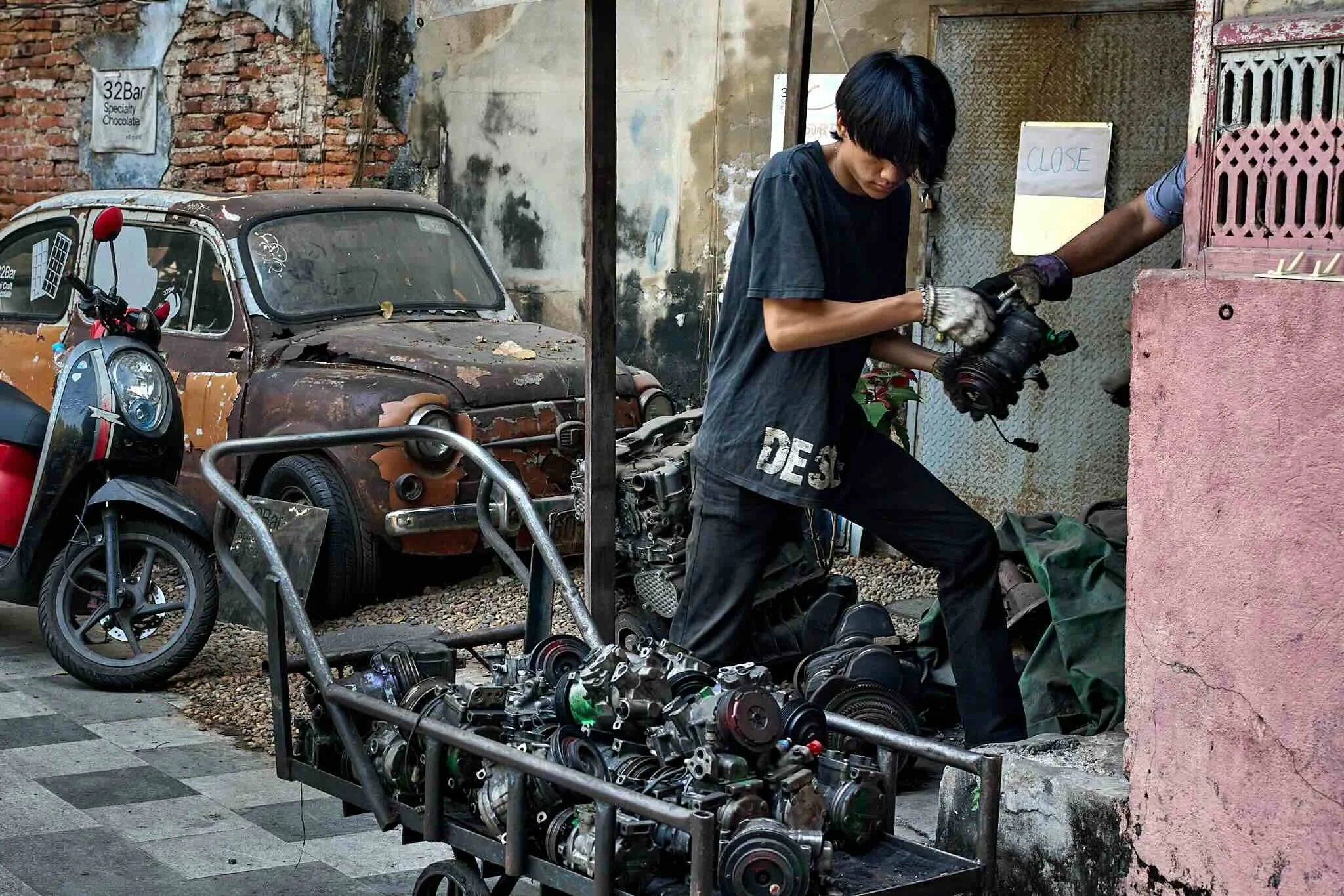 A young man with black hair working on disassembled motorcycle parts outside, near a rusty car and a scooter, in front of an aged brick and concrete wall with a sign that says '32Bar Specialty Chocolate' and another that says 'CLOSE'.