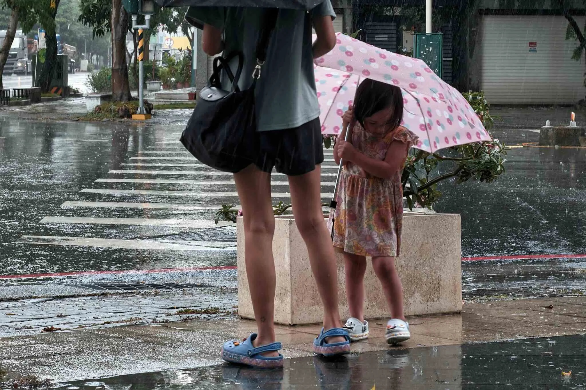 A young girl holding an umbrella standing next to an adult on a rainy city street, with rain pouring and wet pavement reflecting light. The child is wearing a dress and sneakers, and the adult is wearing slides and shorts. There are trees, a parked m