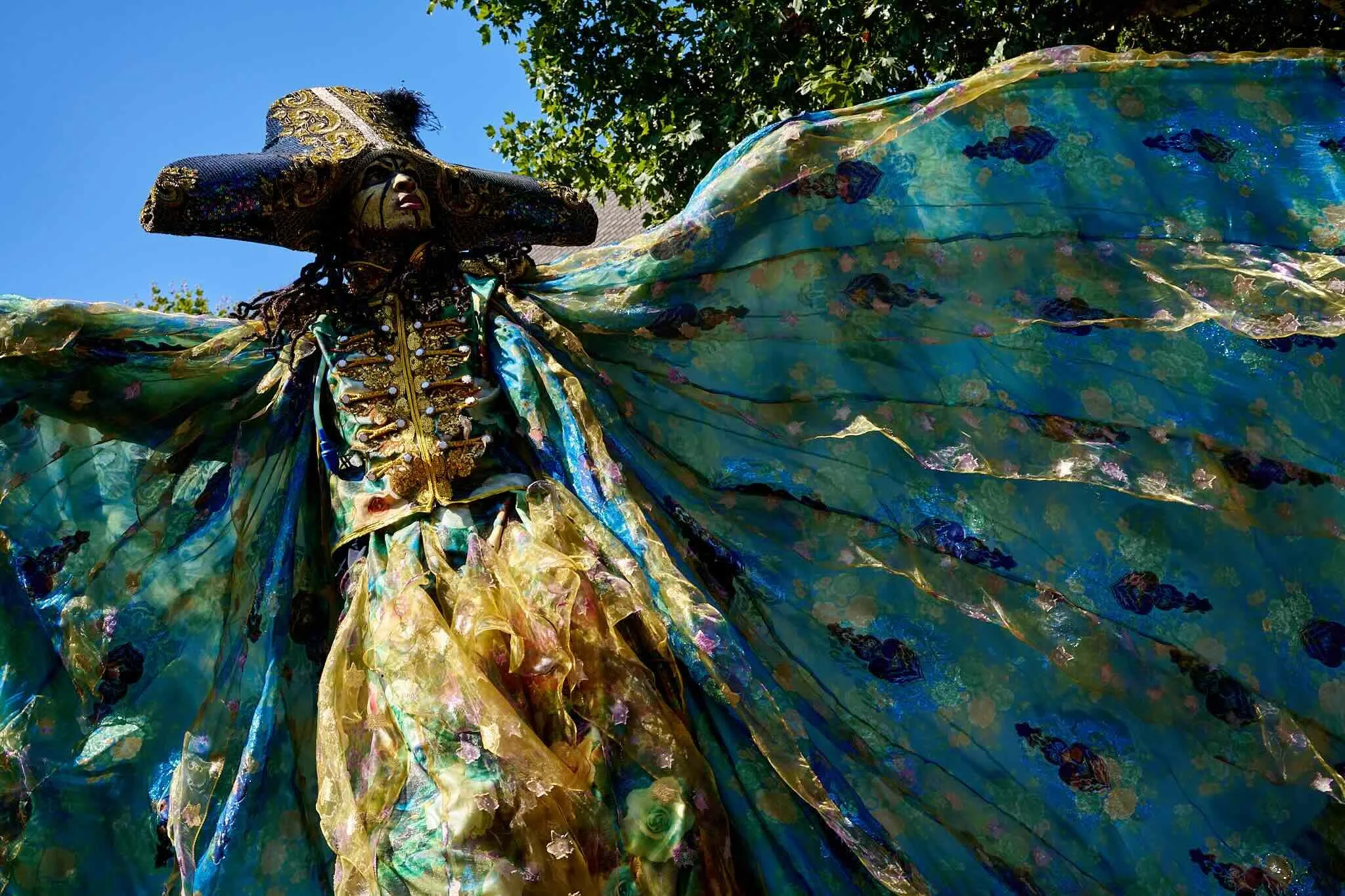 A person wearing an elaborate, colorful costume with a large, decorated hat and flowing fabric, standing outdoors with a clear blue sky and green trees in the background.