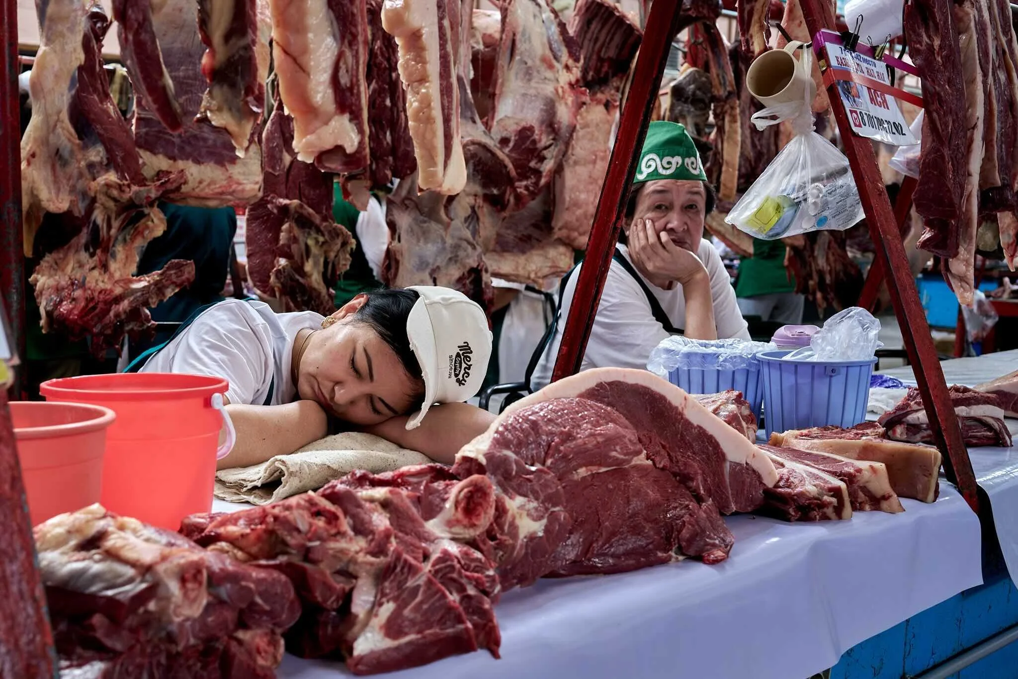 Jerzy Jaworek, Street and Travel Photography, Kazakhstan, Almaty. Market scene with hanging raw meats, including large cuts of beef and other meat, and two women resting behind the stall; one appears to be sleeping, and the other looks contemplative,