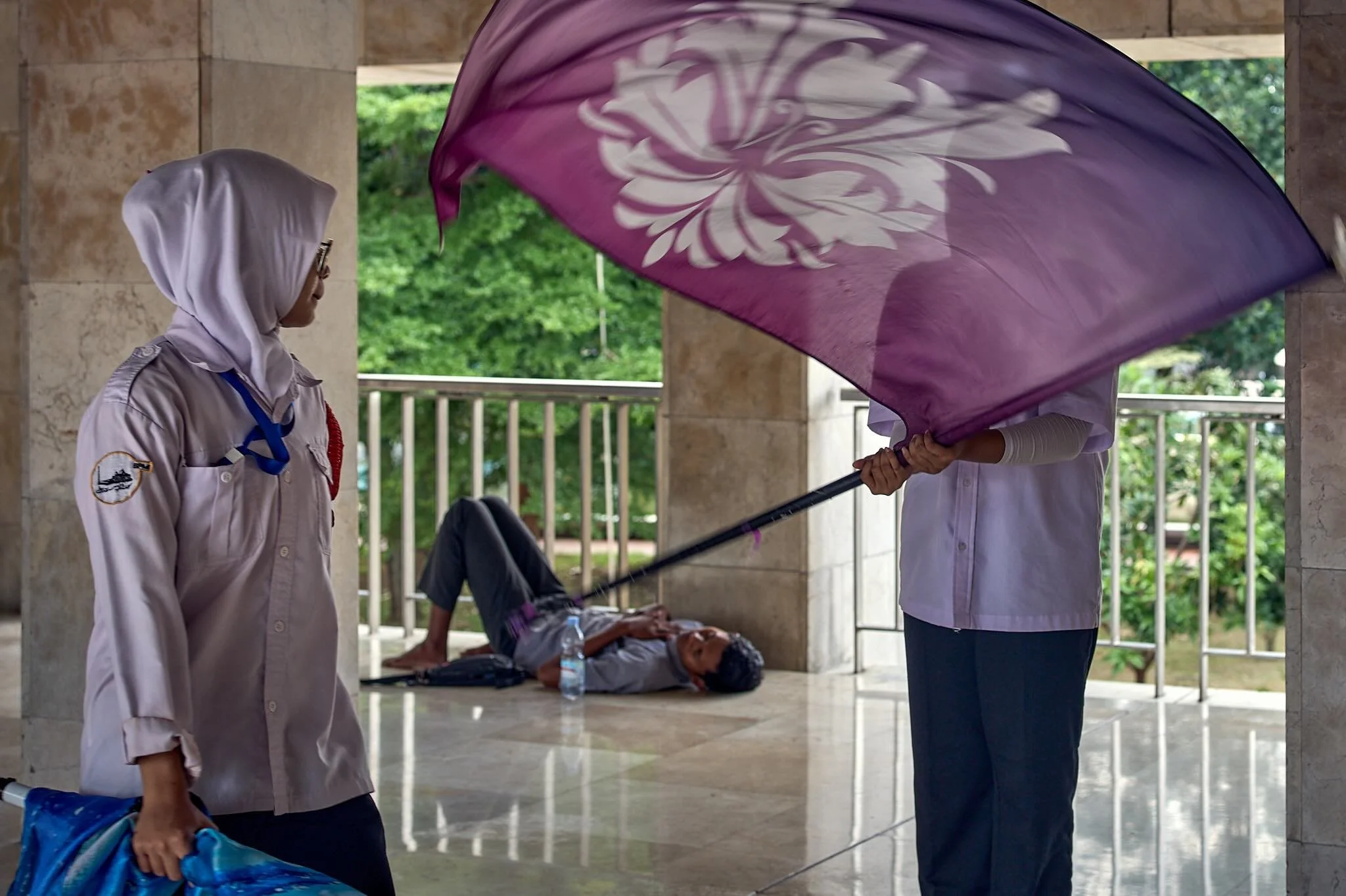 A person lying on the ground under a covered outdoor area, possibly unconscious or resting, with water bottles nearby, while another person dressed in a uniform stands nearby holding a large purple umbrella with a floral design, shading the person.