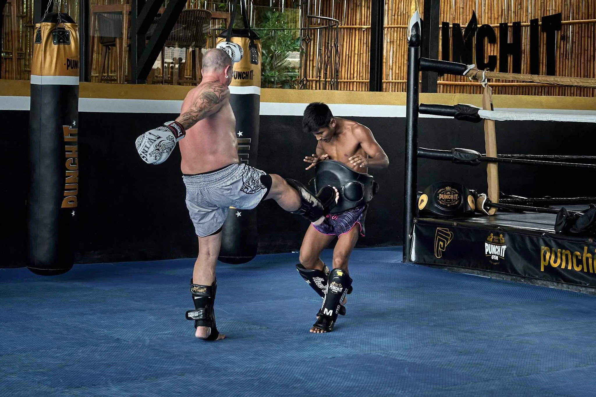 Two shirtless male martial artists practicing kickboxing inside a ring with boxing gloves and shin guards, surrounded by punching bags and training equipment.