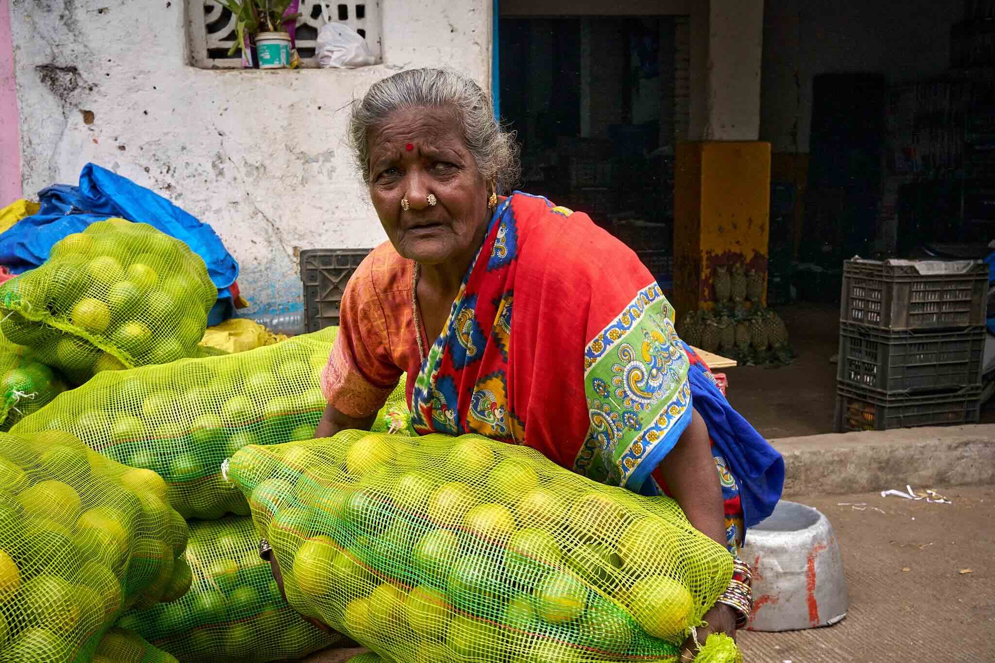 Jerzy Jaworek, Street and Travel Photography, India, Delhi. An elderly Indian woman with gray hair tied back, wearing a colorful sari with red, green, and blue, and gold jewelry, is seated among green mesh bags filled with produce, possibly onions.