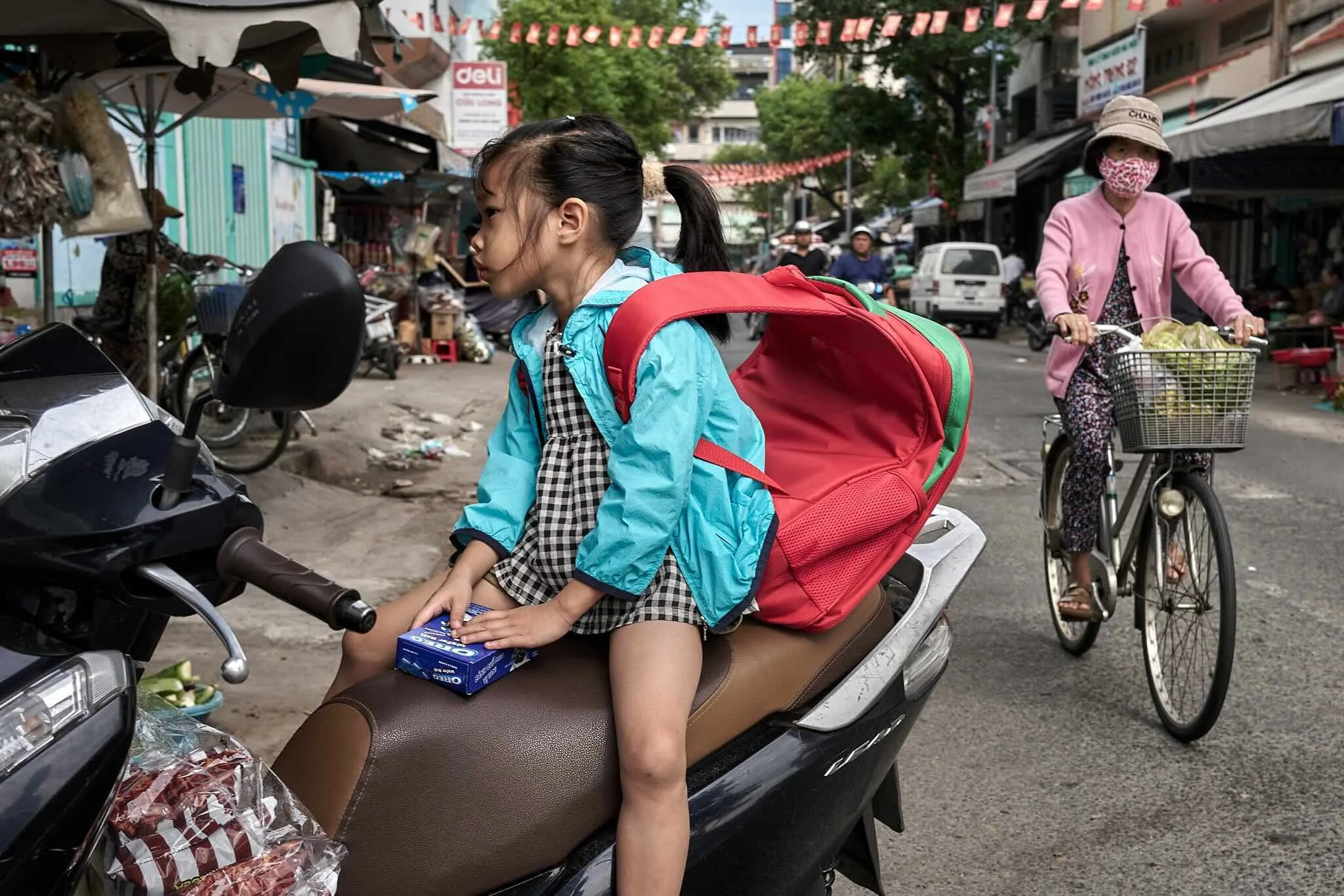 A young girl sitting on a motorcycle on a busy street, looking to the side, with a red and green backpack on her back, holding a box of Oreo cookies. Thai women are riding bicycles in the background, and there are colorful flags and shop signs lining