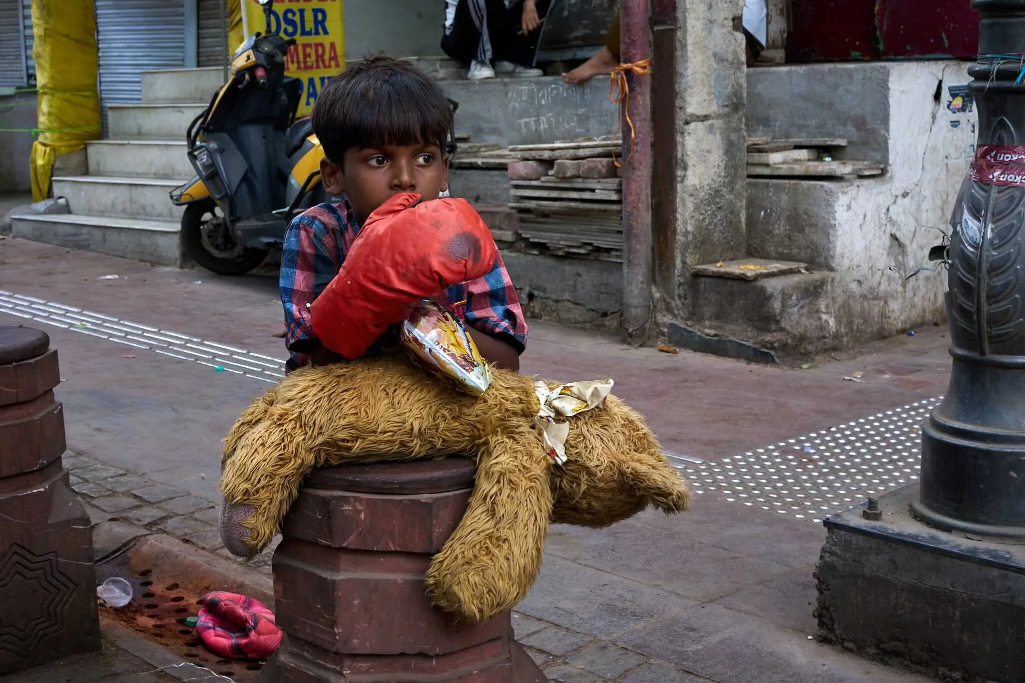 Jerzy Jaworek, Street and Travel Photography, India, Delhi, A young boy sitting on a brick stool on a sidewalk, holding a red bag and surrounded by a large stuffed animal, in an urban setting with stairs, a scooter, and a shop sign in the background.