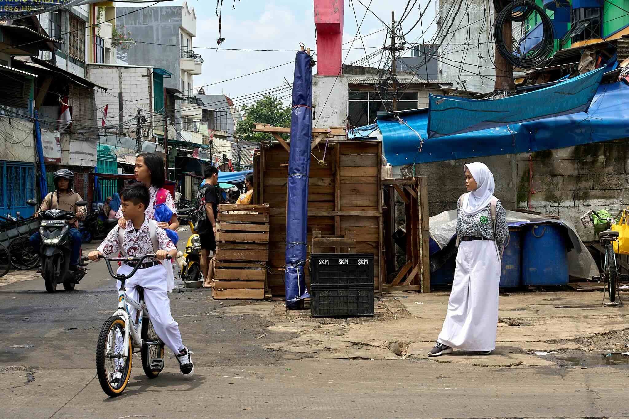 Street scene with pedestrians, children on bikes, motorcycles, and makeshift stalls. Buildings with laundry hanging and electric wires overhead. Some people are dressed in traditional clothing.