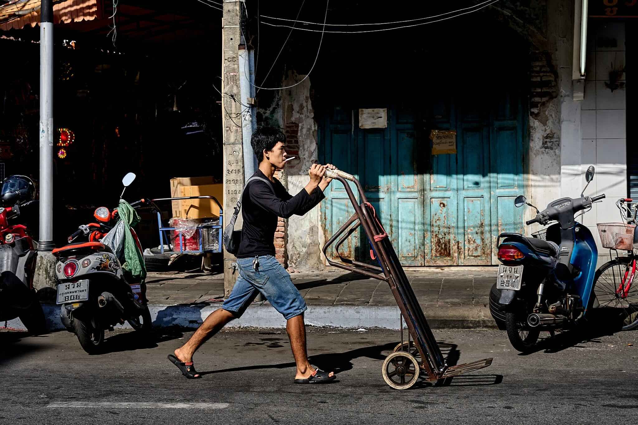 Young man walking barefoot with a cigarette in his mouth, pushing a bicycle on a busy street with parked motorcycles and an old building in the background.