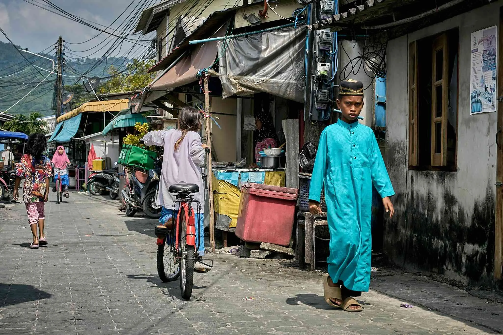 A street scene in a residential area shows children with bicycles and oto r parked motorcycles. A boy in a blue traditional outfit walking on the sidewalk, with houses and power lines in the background.
