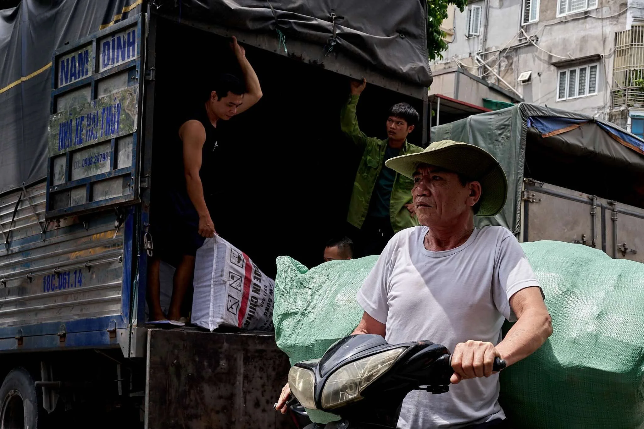 Three men are working around a truck loaded with large bags. One seated man is riding a motorbike on the street in front of the truck. A young man is inside the truck, and another is standing at the back, holding onto the truck’s roof.
