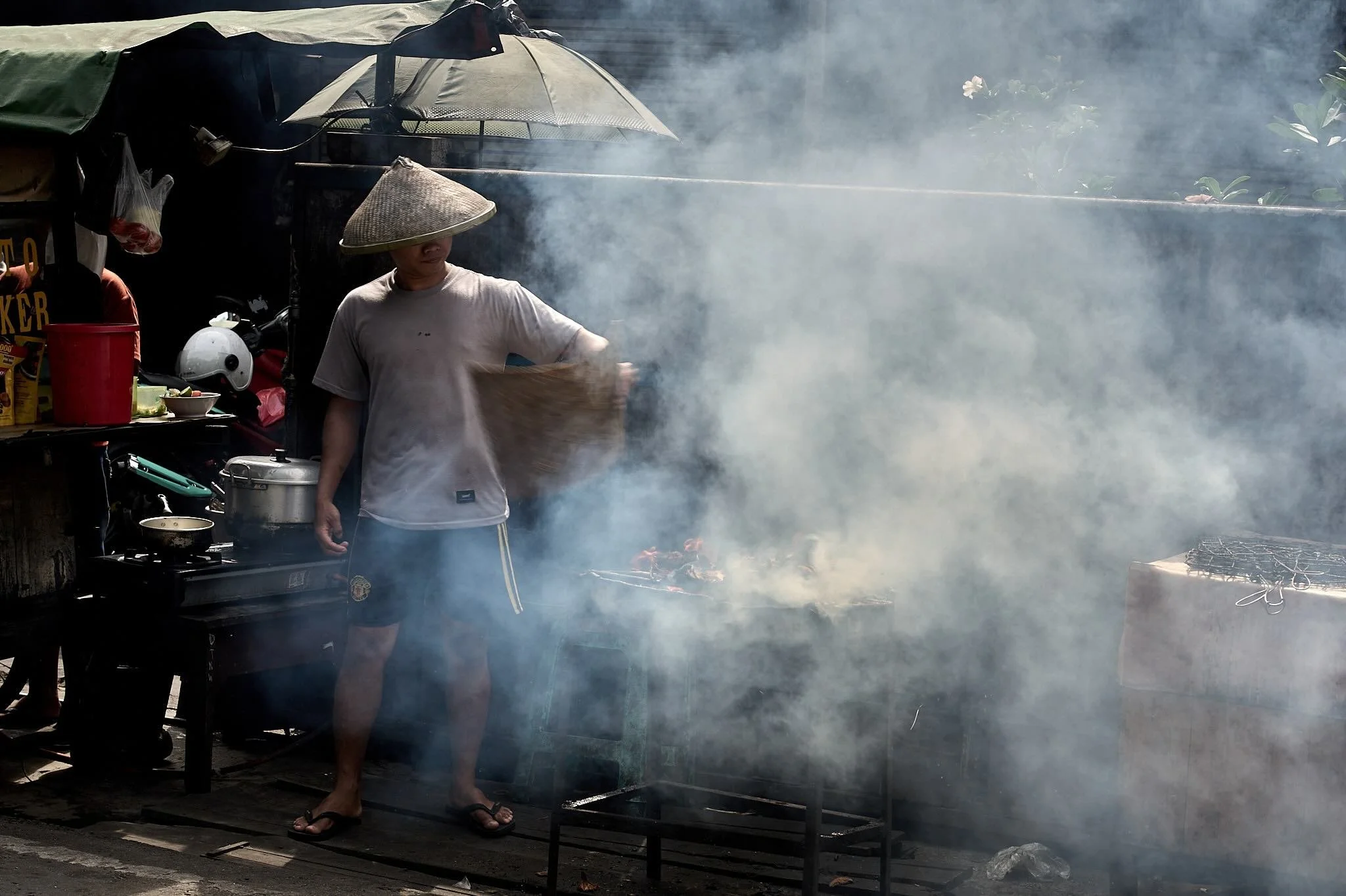 A person wearing a conical hat is cooking outdoors amidst smoke, with a street food stall backdrop including a parasol and various items on a table.