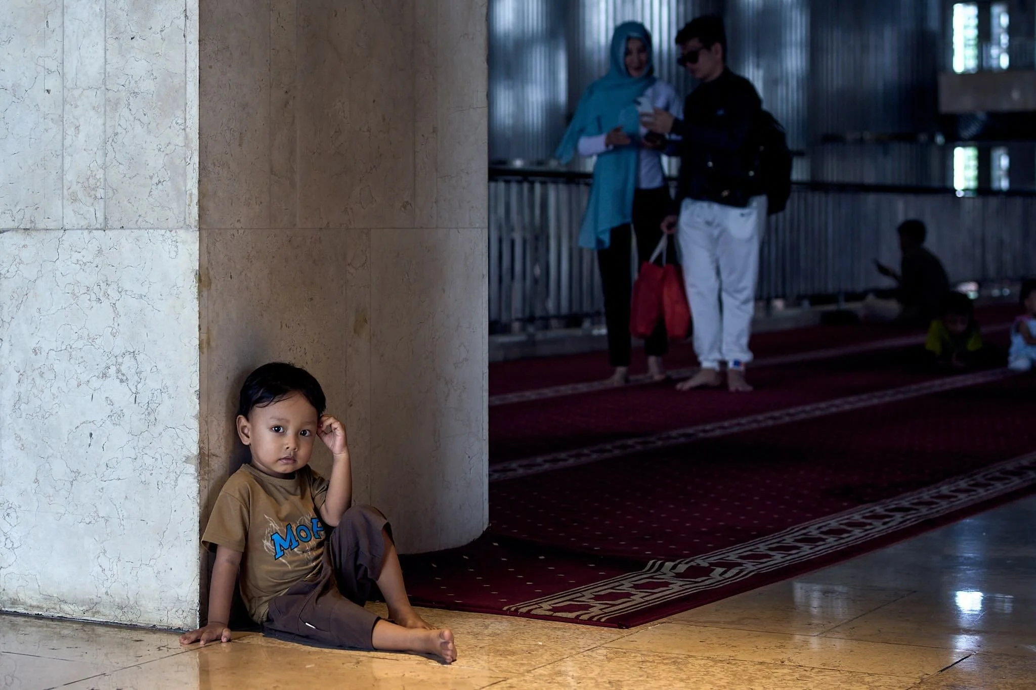 A young boy with dark hair and brown eyes sitting on the floor, leaning against a marble wall, looking at the camera. He is wearing a brown T-shirt and brown pants. Other children can be seen in the background, with some standing and others sitting o