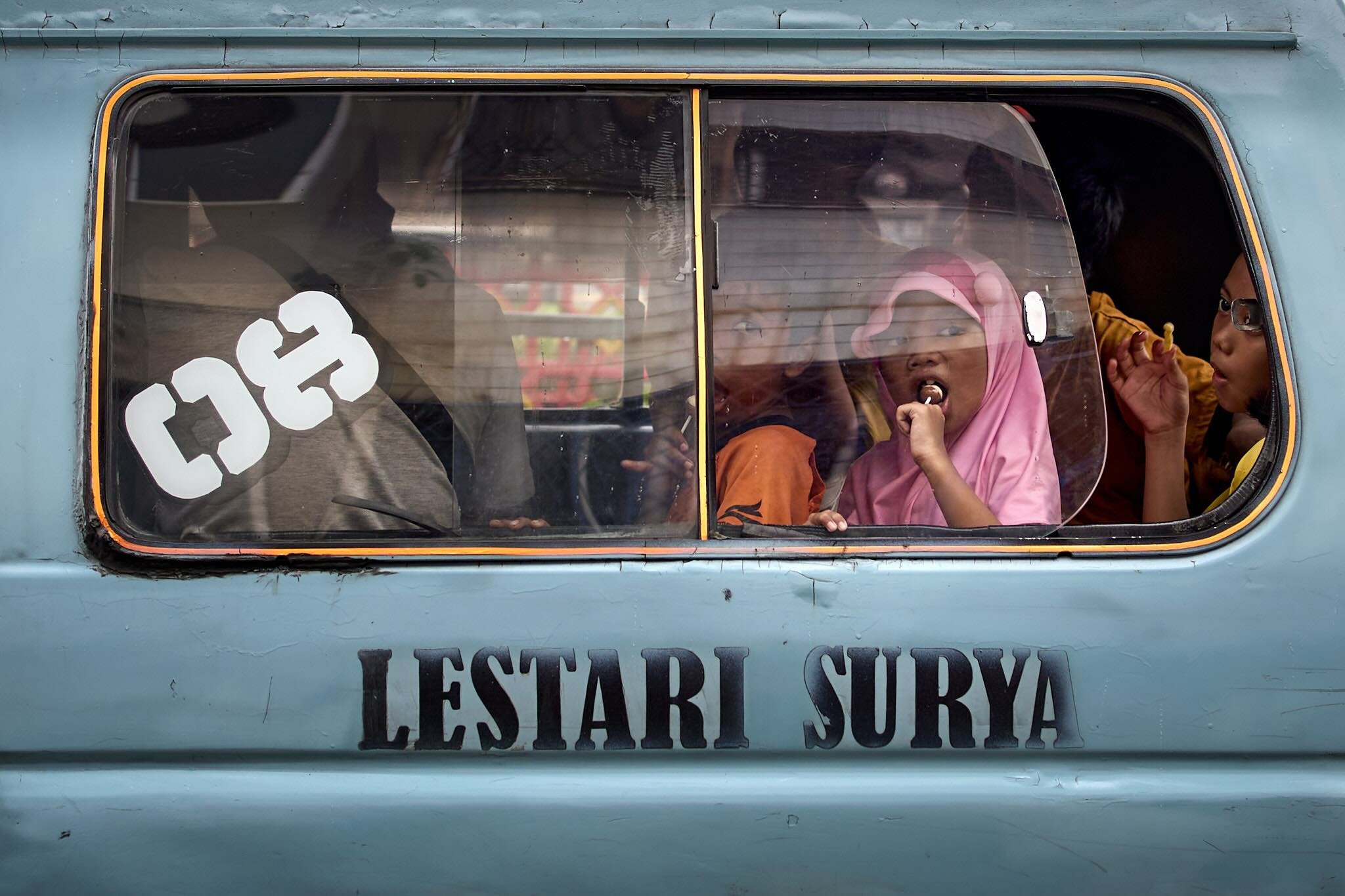 Children sitting inside a blue vehicle, one girl wearing a pink hijab is eating a snack, with other children visible through the window.