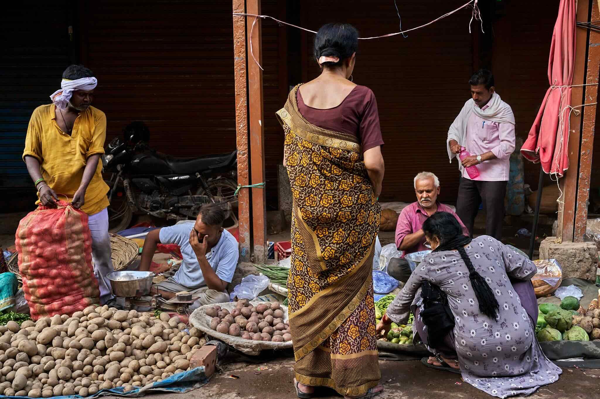 Jerzy Jaworek, Street and Travel Photography, India, Delhi. A group of people at an outdoor market in India, with vendors selling potatoes, vegetables, and other produce, some sitting and some standing.