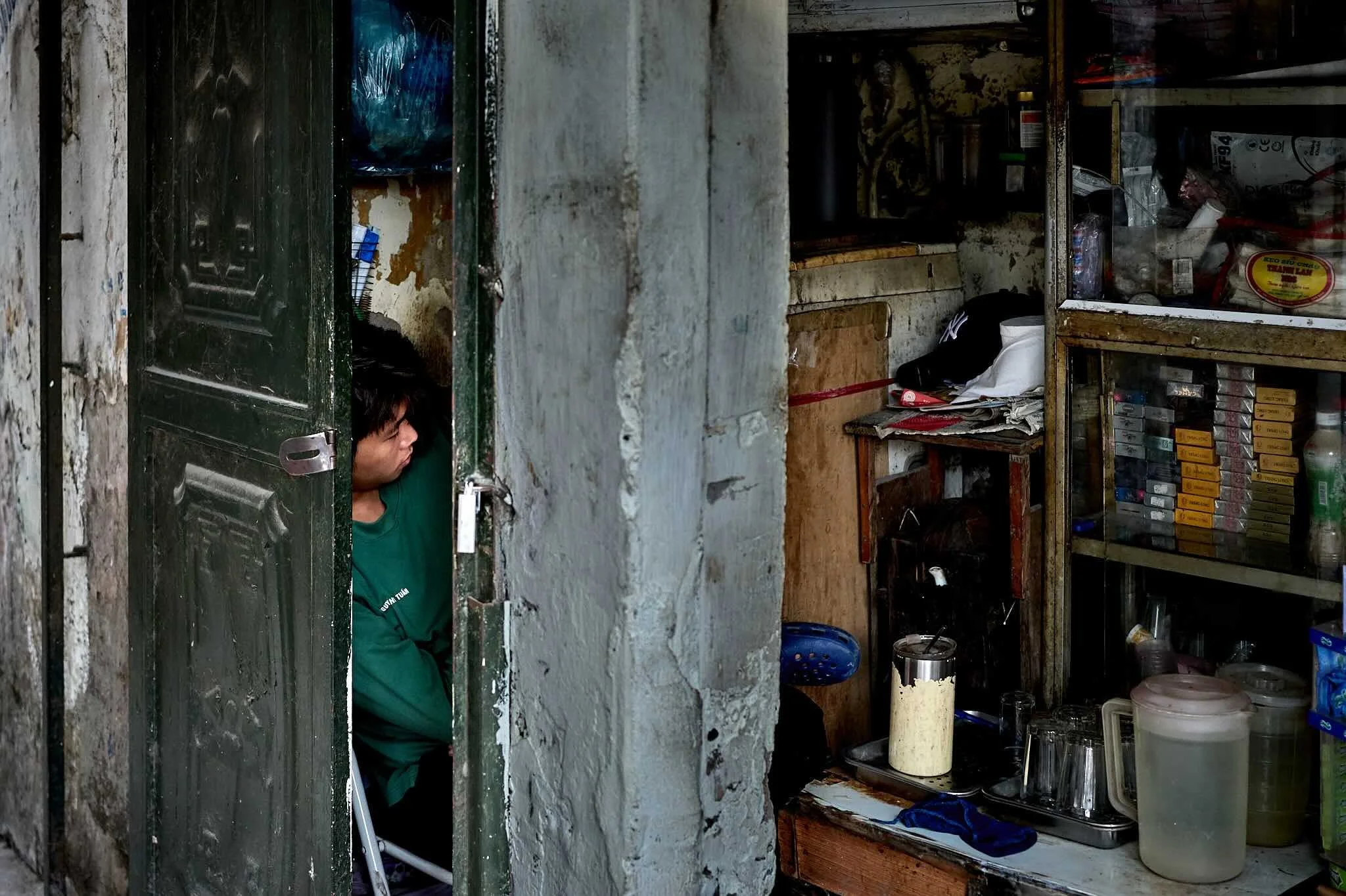 A small store with a man wearing a green shirt behind the metal grill serving as a window. Inside, shelves are filled with various household and food items, including jars, boxes, and containers.