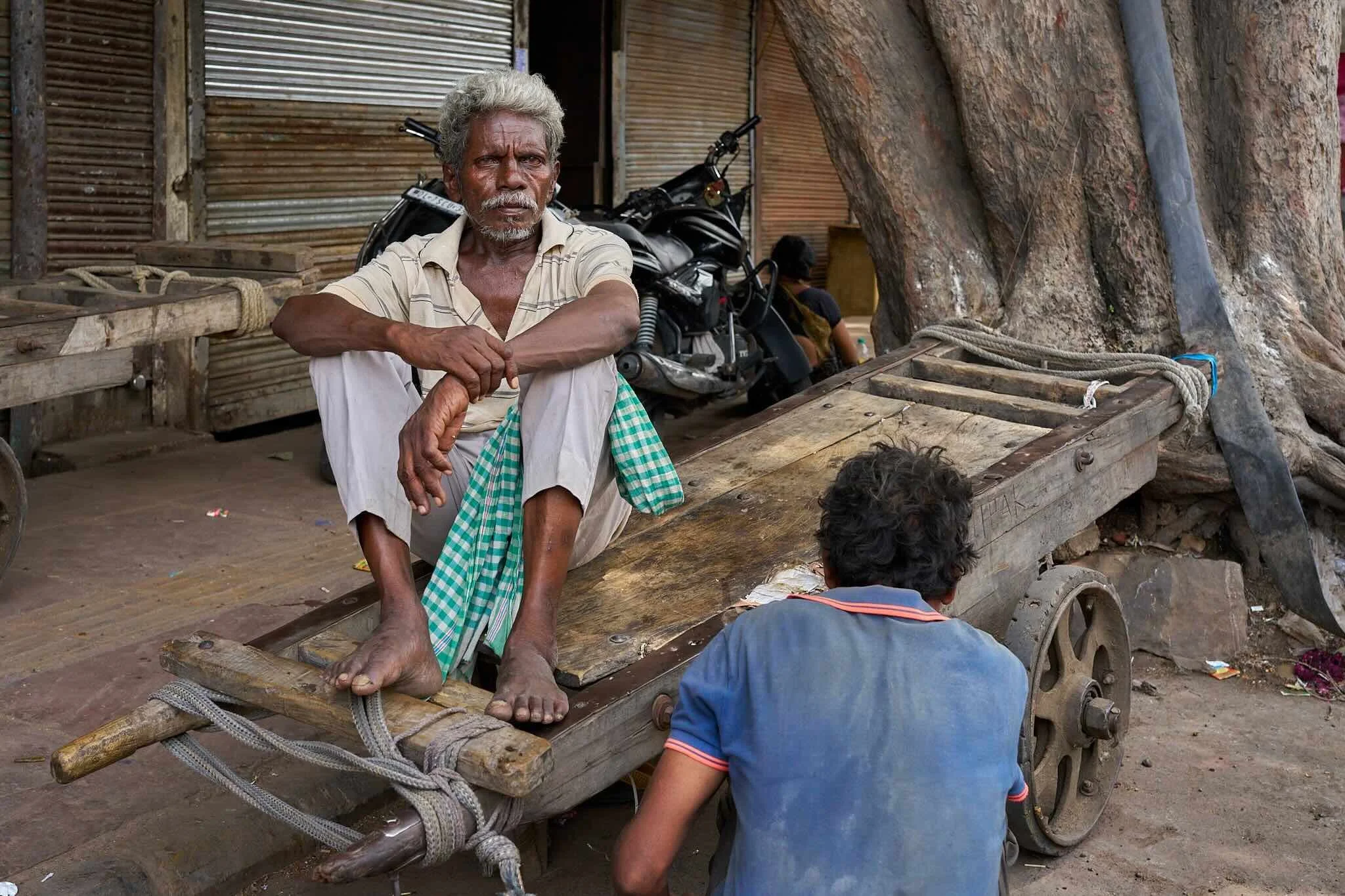 Jerzy Jaworek, Street and Travel Photography, India, Delhi. An elderly man with gray hair and a mustache sitting on a wooden cart in front of a rustic building with metal and wooden walls, a motorcycle, and children nearby.