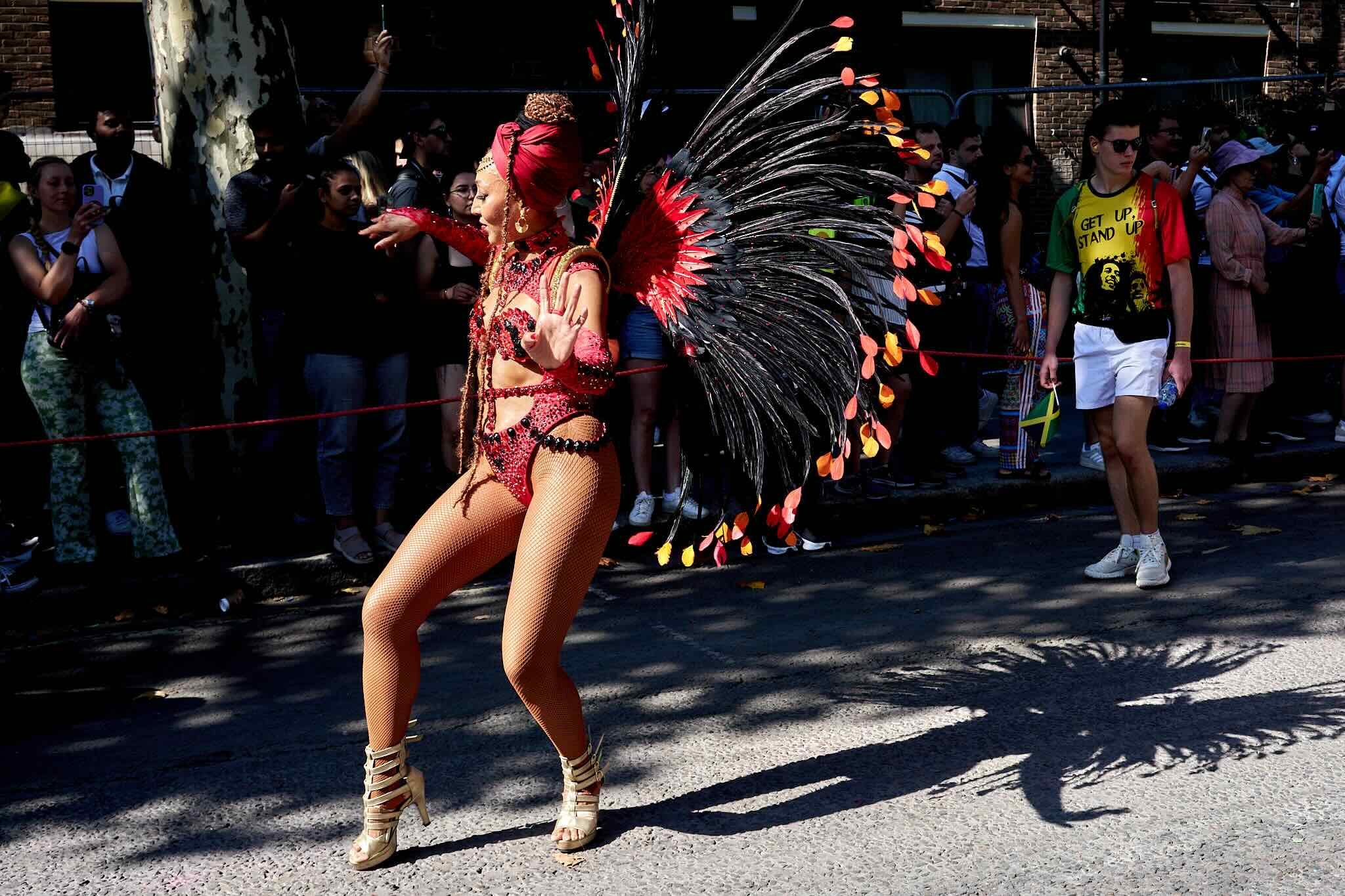 A person in an elaborate red and black feathered costume with large wings and fishnet tights is dancing in front of a crowd on a city street during a parade.