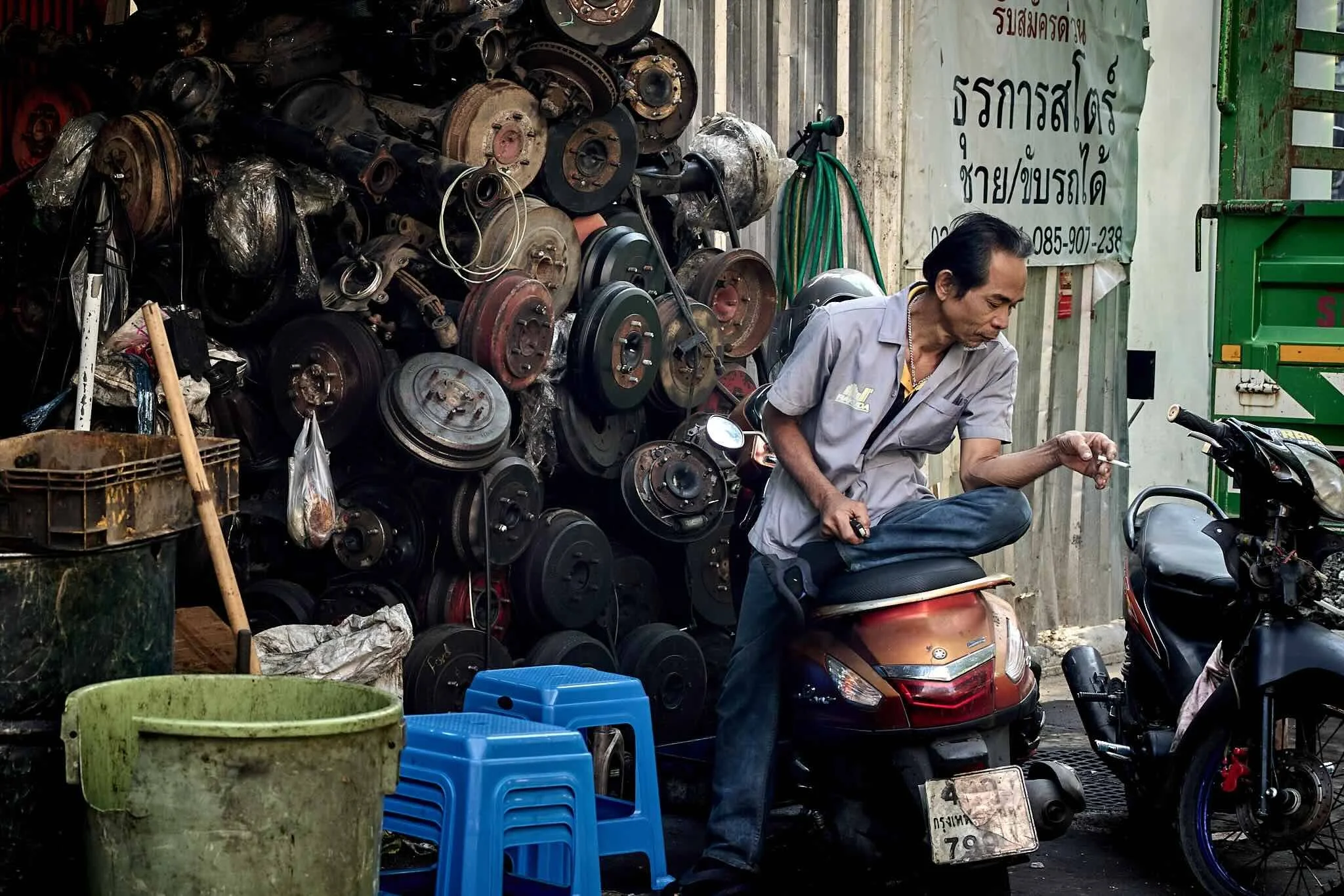 A man sitting on a motorcycle in front of a pile of used car engine parts, with plastic chairs, buckets, and a scooter visible nearby.