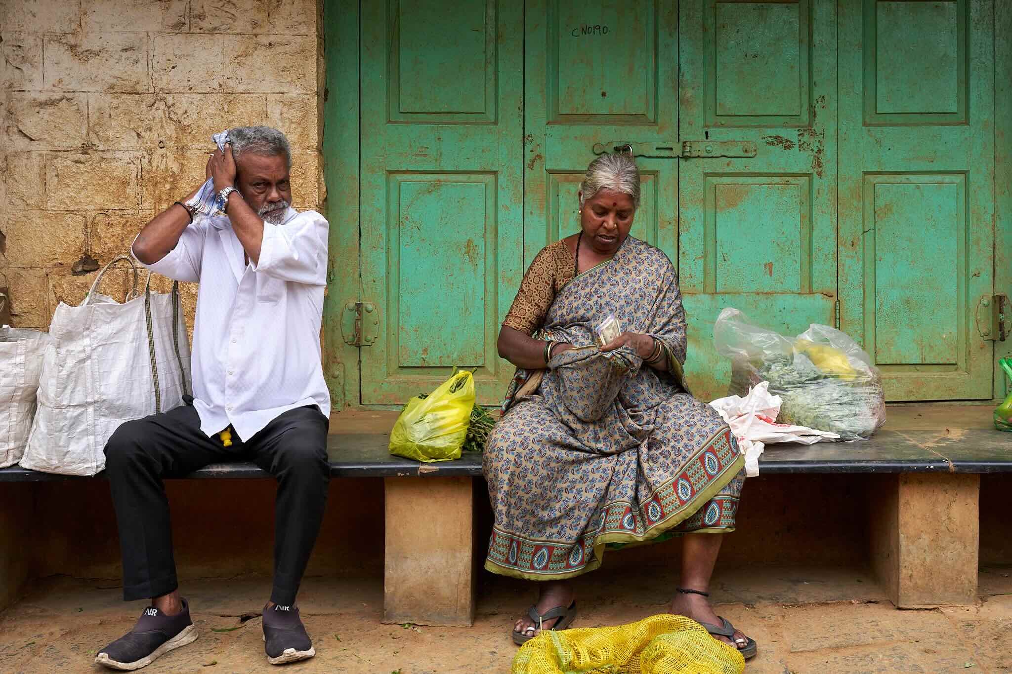 Jerzy Jaworek, Street and Travel Photography, India, Delhi. An older man and woman sitting on a ledge against a green door with closed drawers. The man, with gray hair and a beard, is holding a piece of cloth to his head.