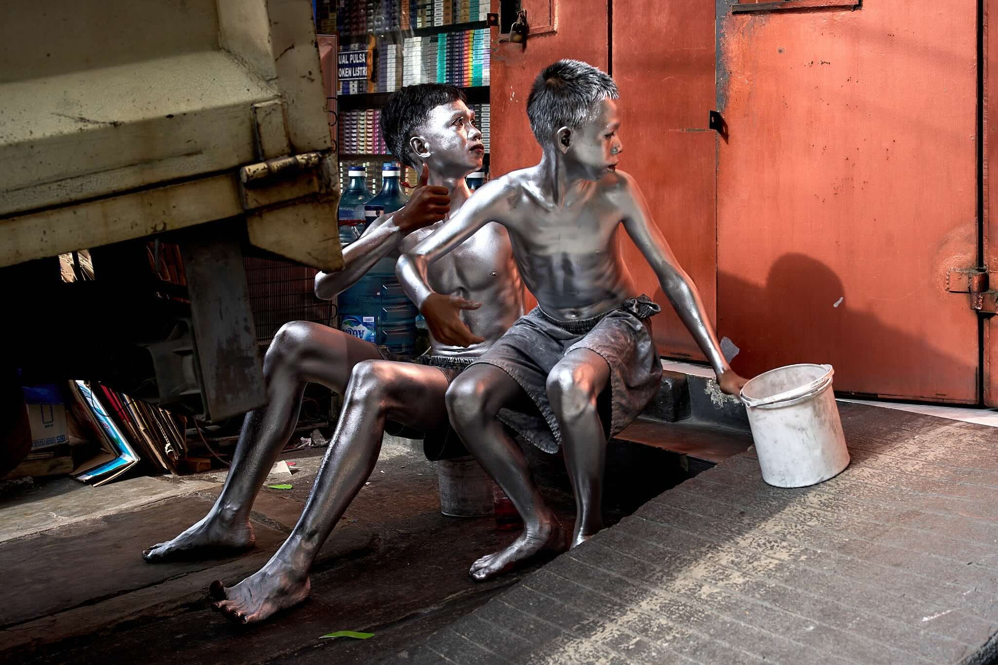 Two life-sized statues of children sitting on a bench, painted in a metallic silver color, in a shop with shelves of books in the background and large water bottles behind them.