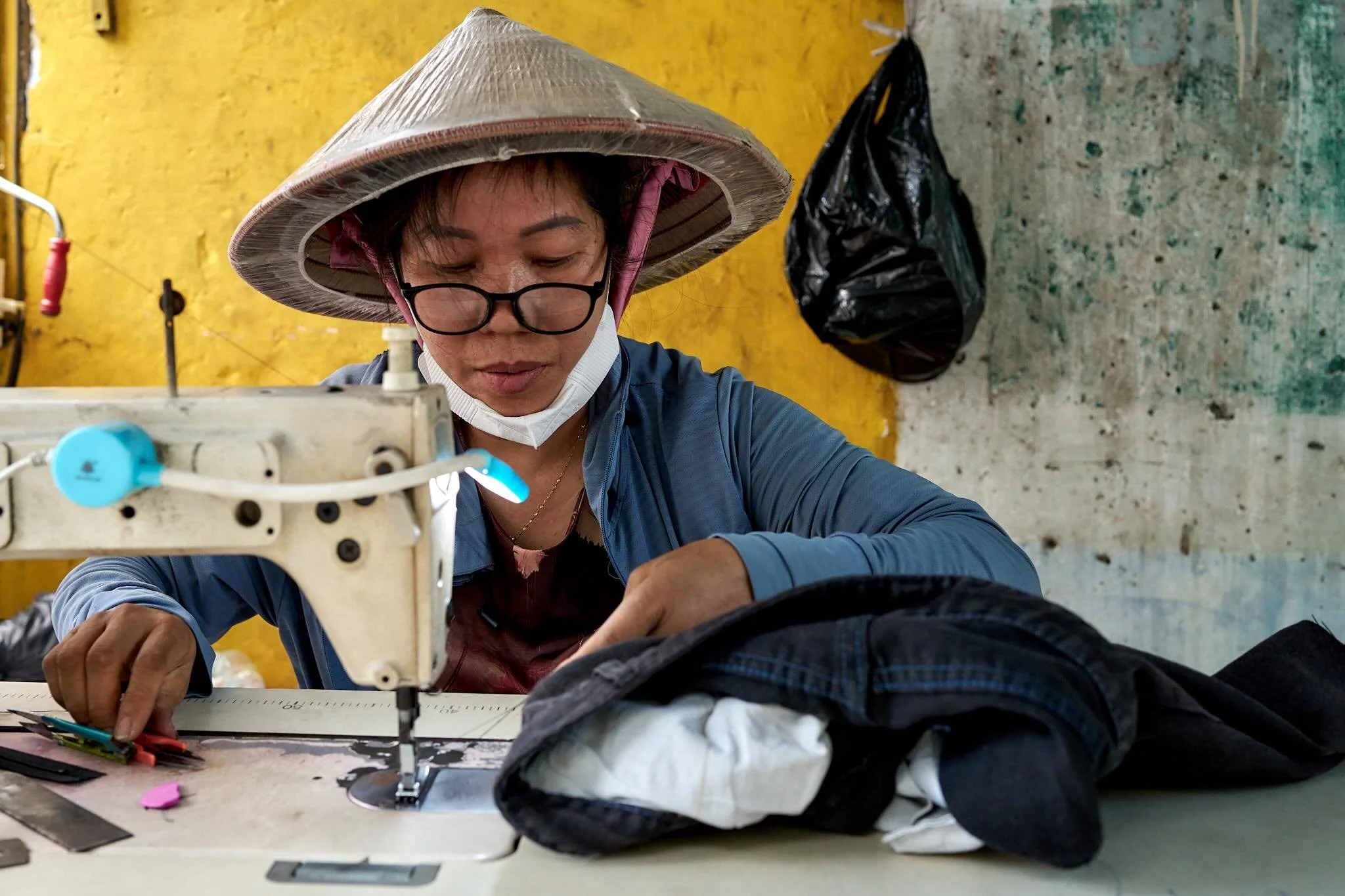 A woman wearing glasses, a conical hat, a face mask, and a blue jacket working on a piece of fabric or clothing at a sewing machine in a yellow-walled workshop.