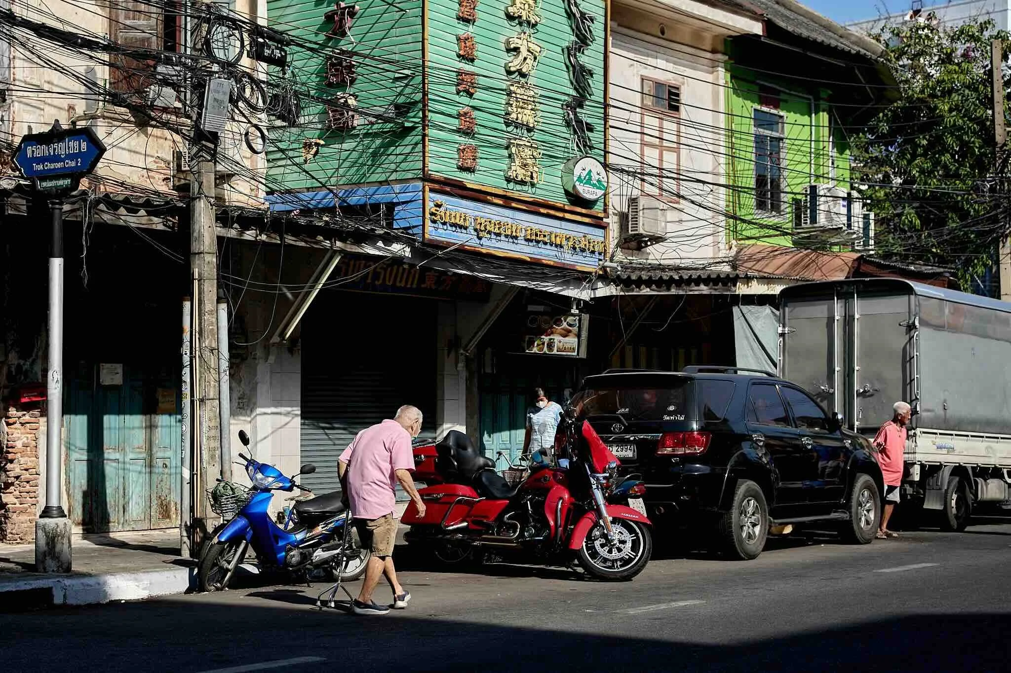 Street scene with motorcycles, cars, and pedestrians in front of a building with Thai signs, electrical wires, and colorful facades.