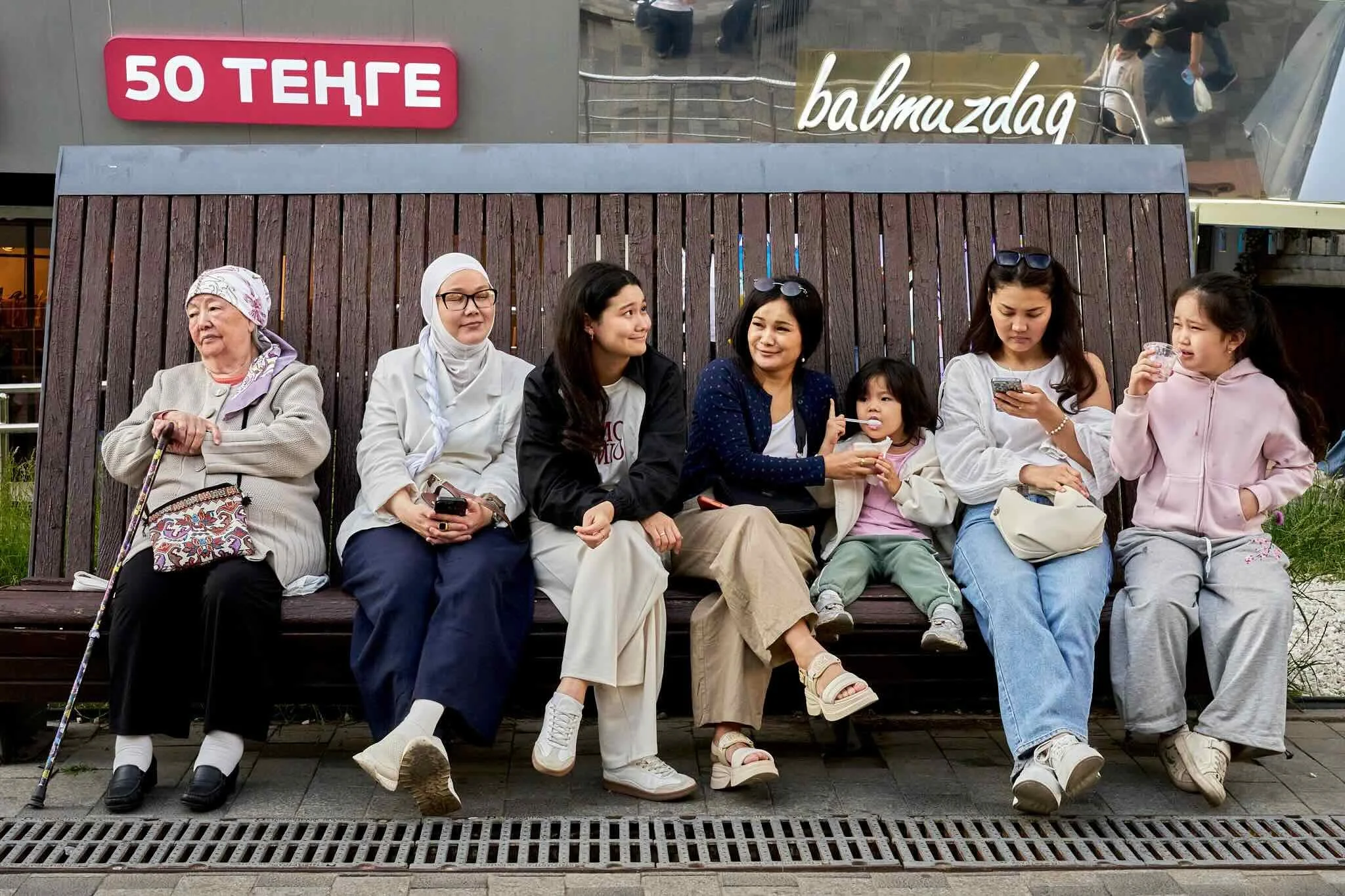 Jerzy Jaworek, Street and Travel Photography, Kazakhstan, Almaty. A diverse group of women and girls sitting on a wooden park bench outside, some looking at their phones and others conversing, with a building with signs behind them.