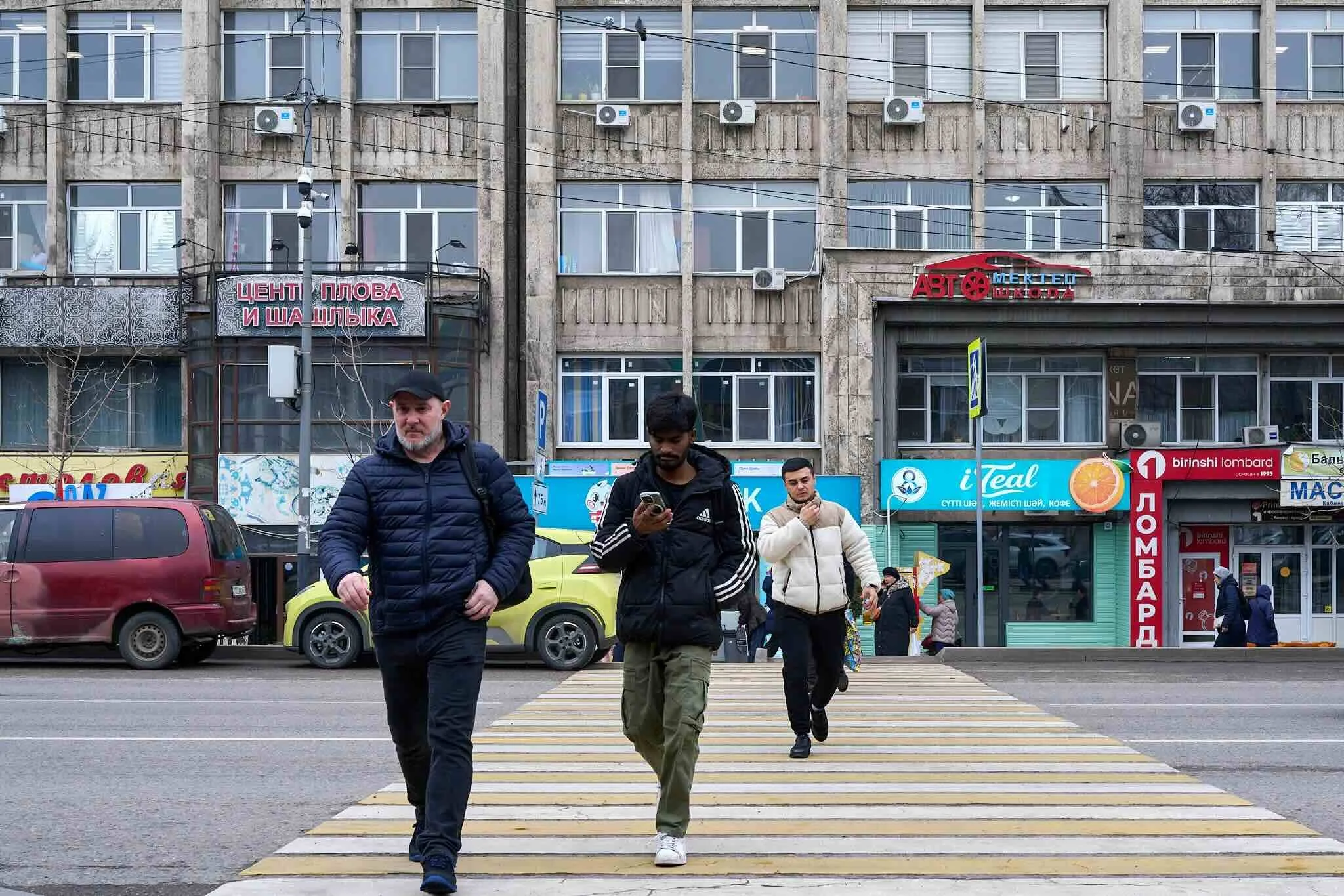 Jerzy Jaworek, Street and Travel Photography, Kazakhstan, Almaty. People walking across a yellow pedestrian crosswalk in front of a building with shop signs.