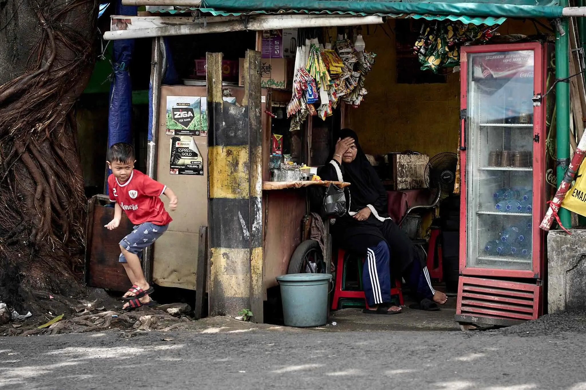 A small street-side shop with a woman and a young boy. The woman is sitting inside, holding her head, with various items like snack packets hanging and placed around her. The boy in a red shirt is standing outside, looking to the side. The shop has a