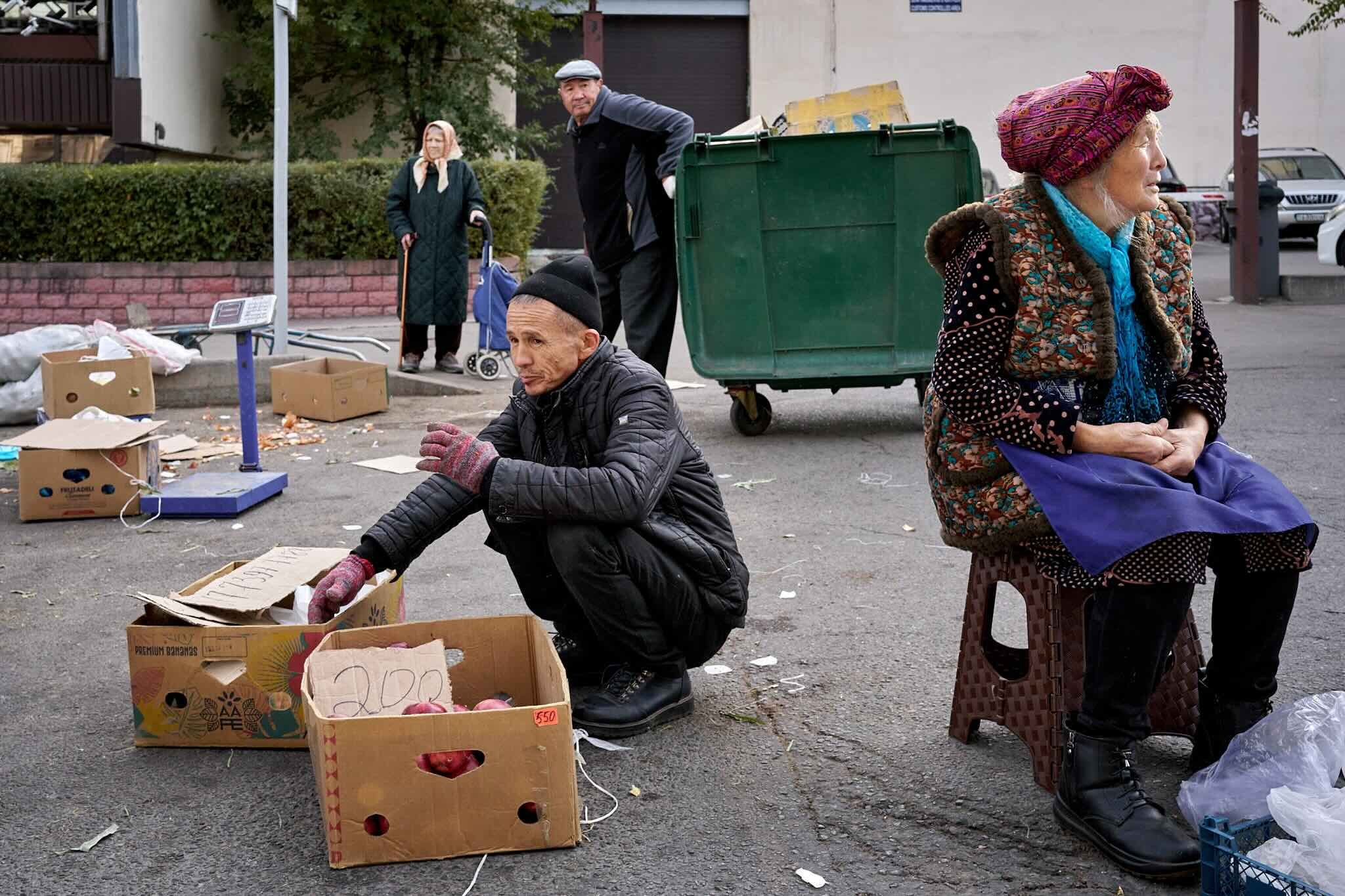 Jerzy Jaworek, Street and Travel Photography, Kazakhstan, Almaty. Several elderly people gathered outside on the street near cardboard boxes filled with produce, with some sitting and some standing. There is a large green trash bin in the background,