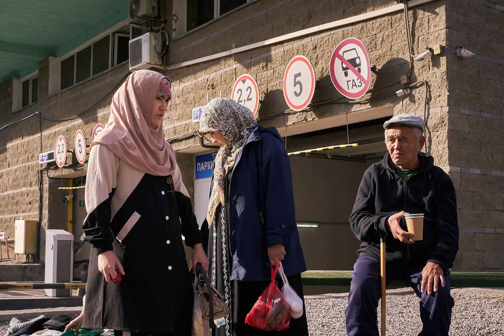 Jerzy Jaworek, Street and Travel Photography, Kazakhstan, Almaty. Three people waiting in line outside a building with parking signs, including no gas and speed limit signs, in the background.
