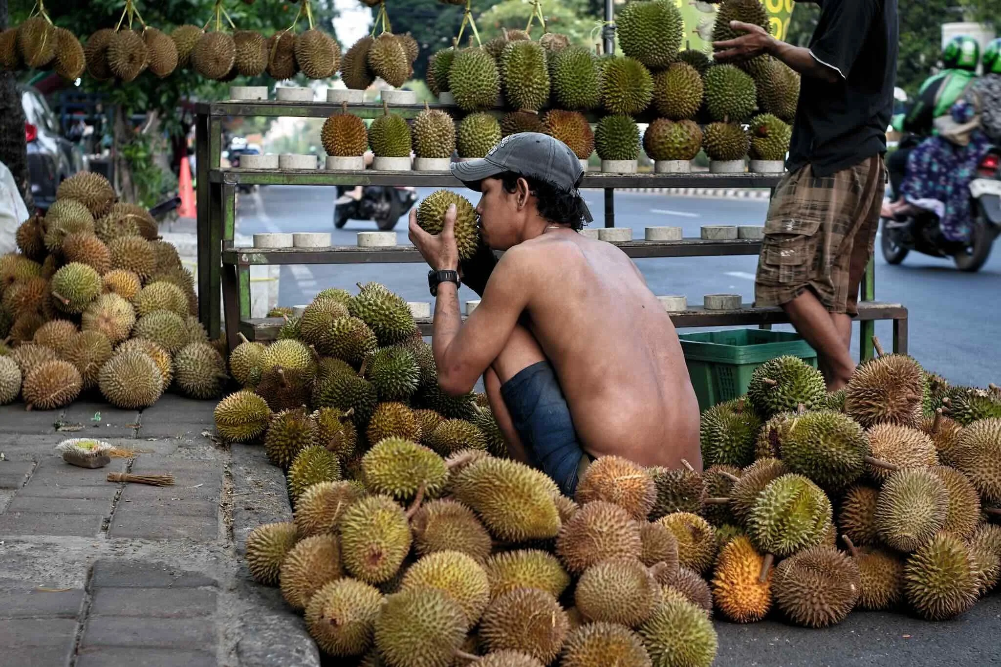 A man with no shirt and a cap sniffs a durian fruit at an outdoor durian stand, with durians on display and people in the background.