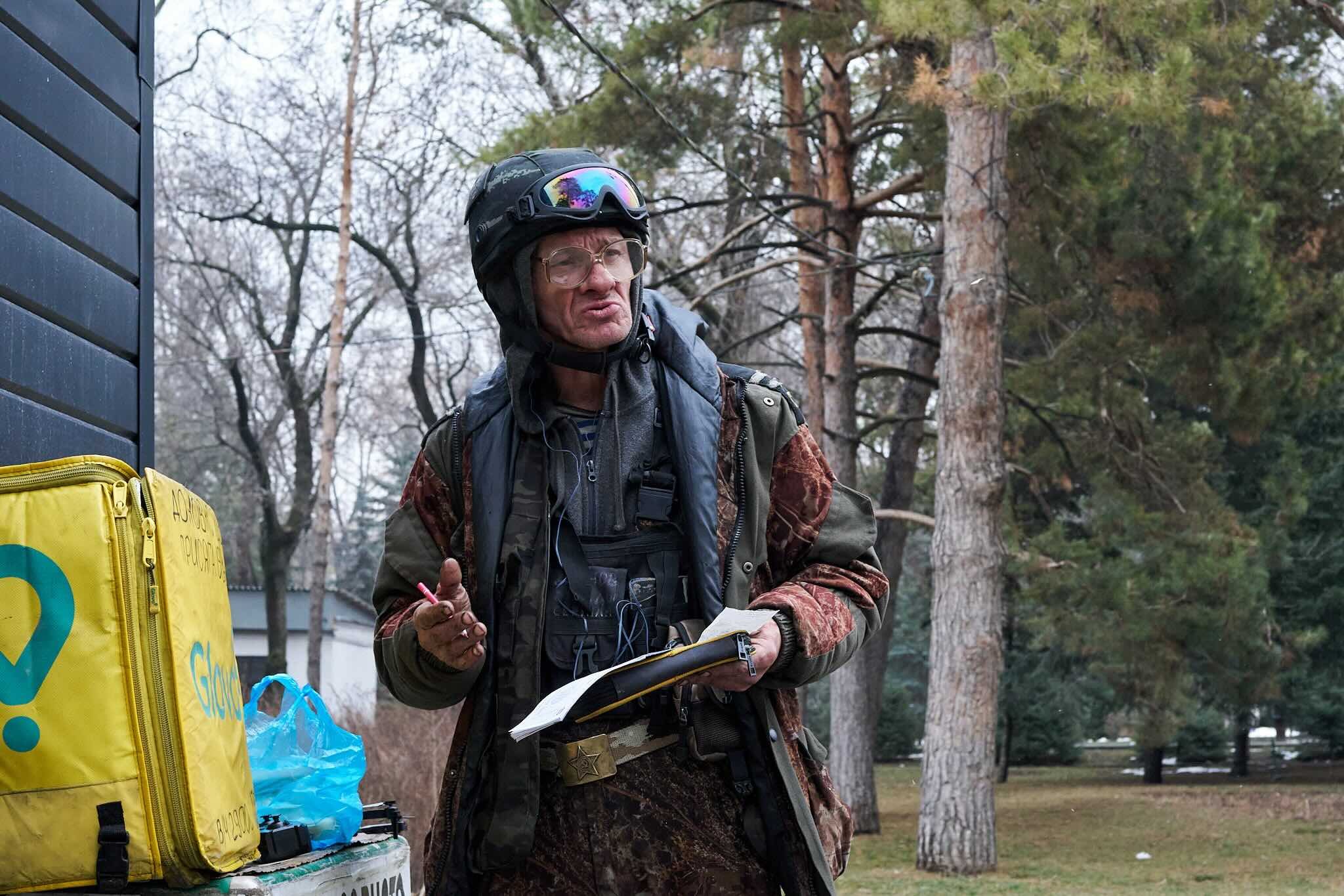 Jerzy Jaworek, Street and Travel Photography, Kazakhstan, Almaty. A man wearing a helmet with goggles, glasses, a backpack, and camouflage clothing, holding a pen and notepad, standing outdoors among trees.