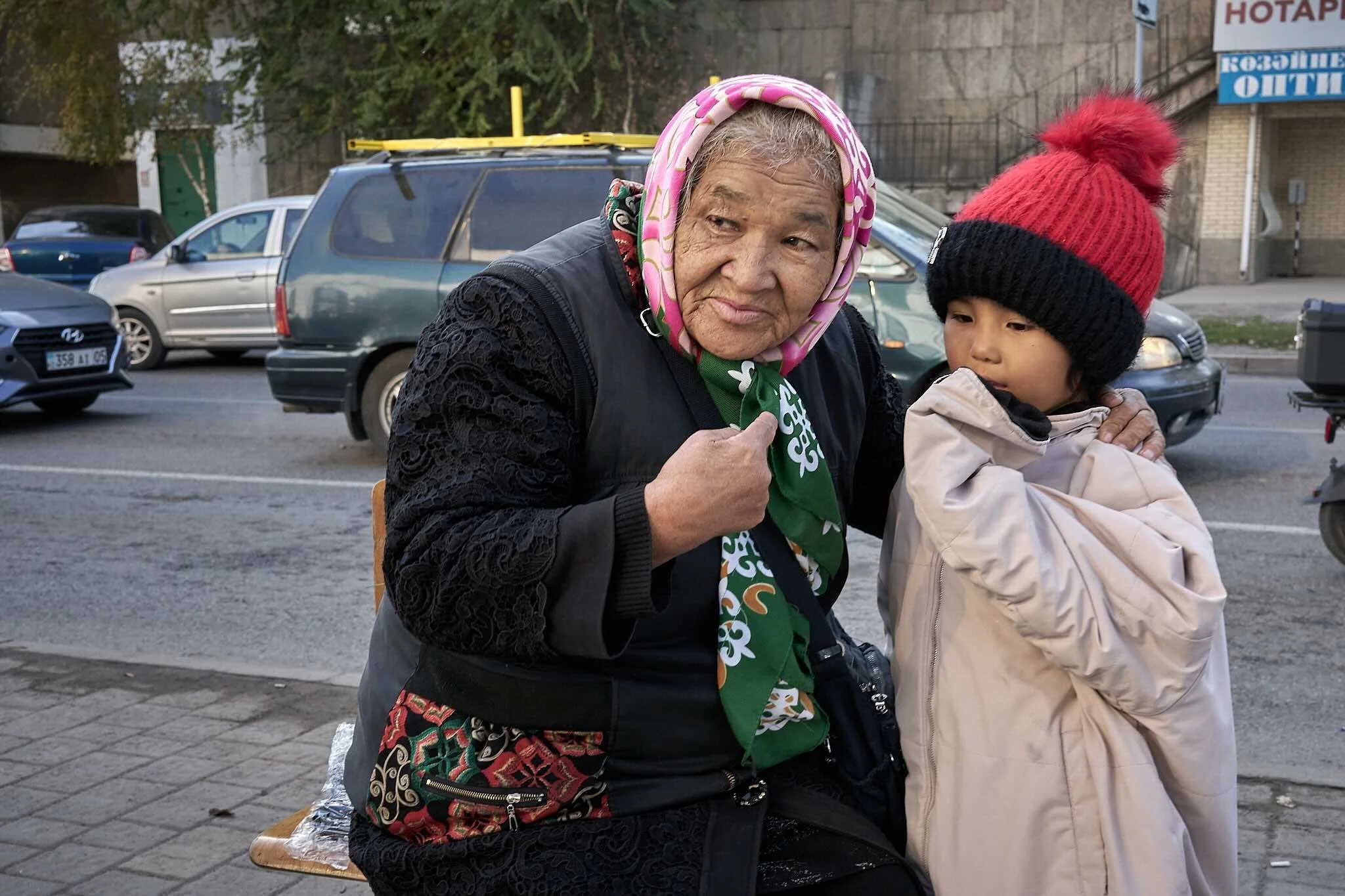 Jerzy Jaworek, Street and Travel Photography, Kazakhstan, Almaty. An elderly woman wearing a pink headscarf and black lace jacket sits on a bench next to a young girl wearing a red knit hat with a pom-pom, in a parking lot with cars and buildings in 