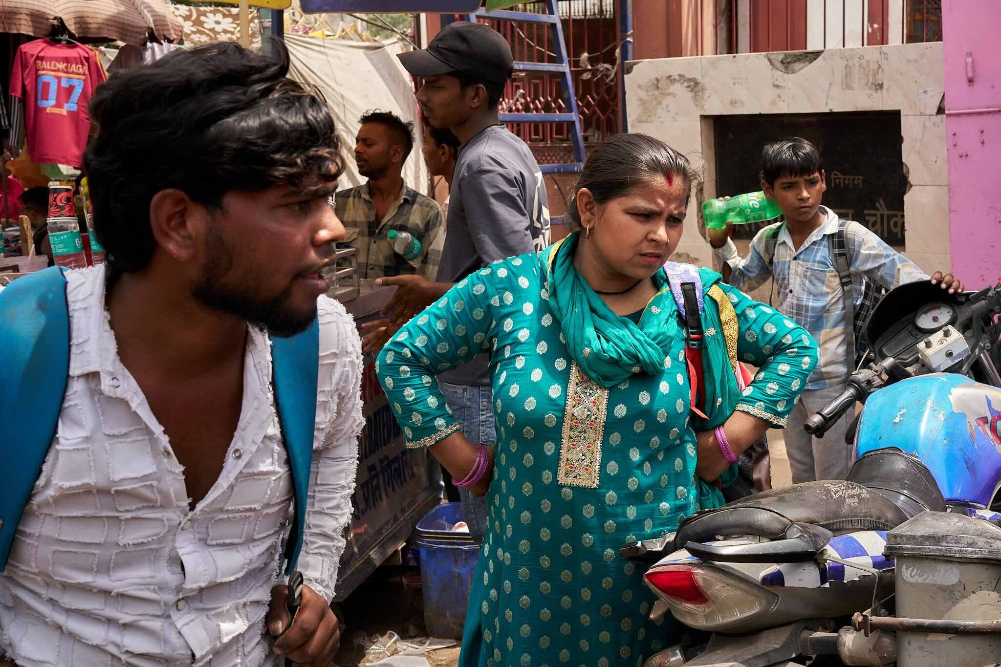Jerzy Jaworek, Street and Travel Photography, India, Delhi, A group of people, including a woman in a blue traditional Indian dress, are gathered around motorcycles on a busy street with shops and hanging clothing in the background.
