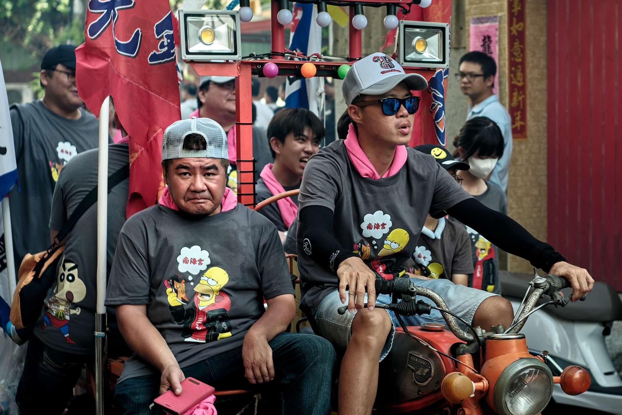 A group of people riding a decorated bicycle or rickshaw during a festival or parade, with some wearing T-shirts featuring a cartoon character.