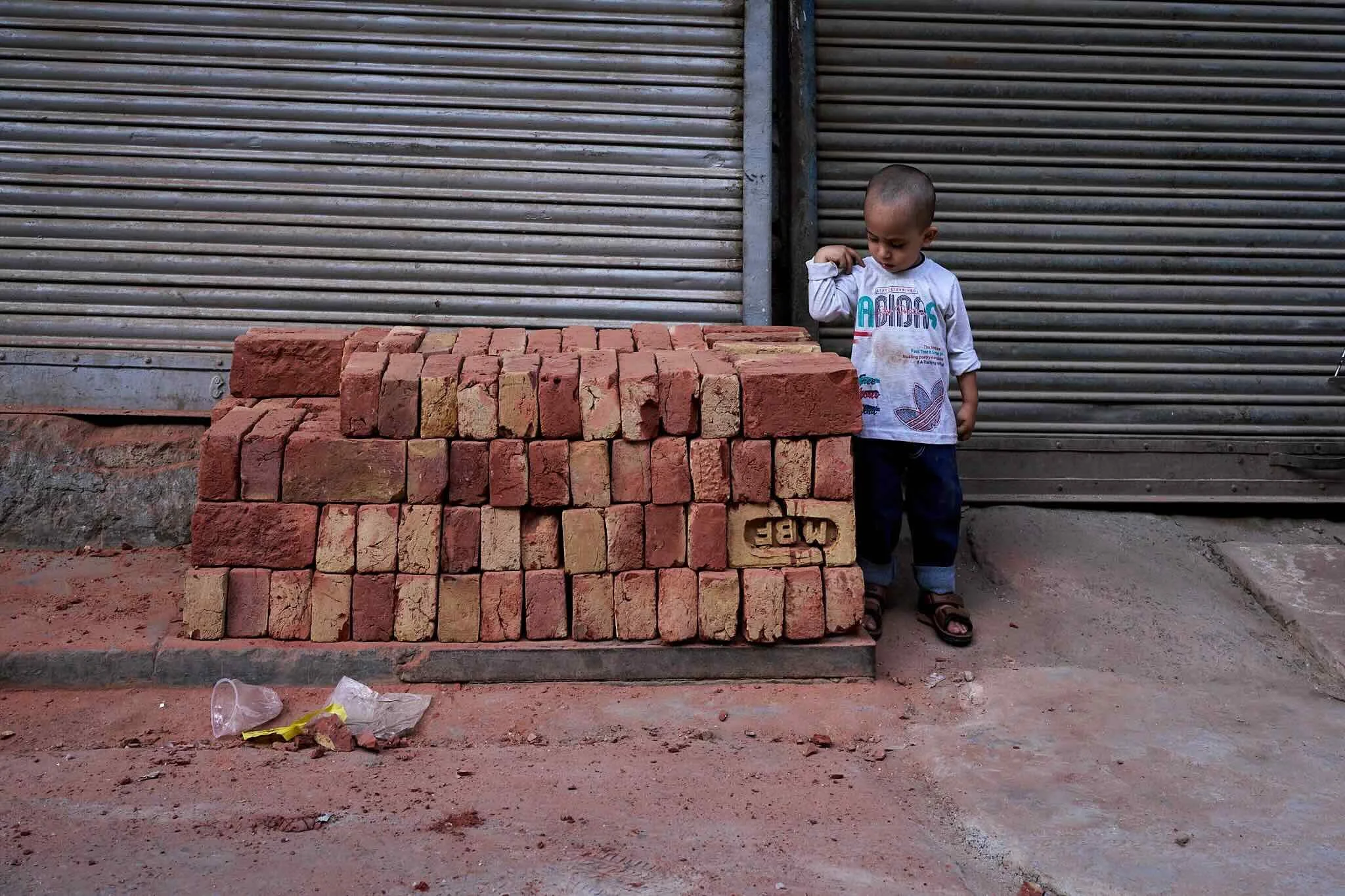 Jerzy Jaworek, Street and Travel Photography, India, Delhi, A young boy in a white Adidas shirt stands next to a large stack of red bricks, against a metal rolling door background, on a dirt ground with some trash.