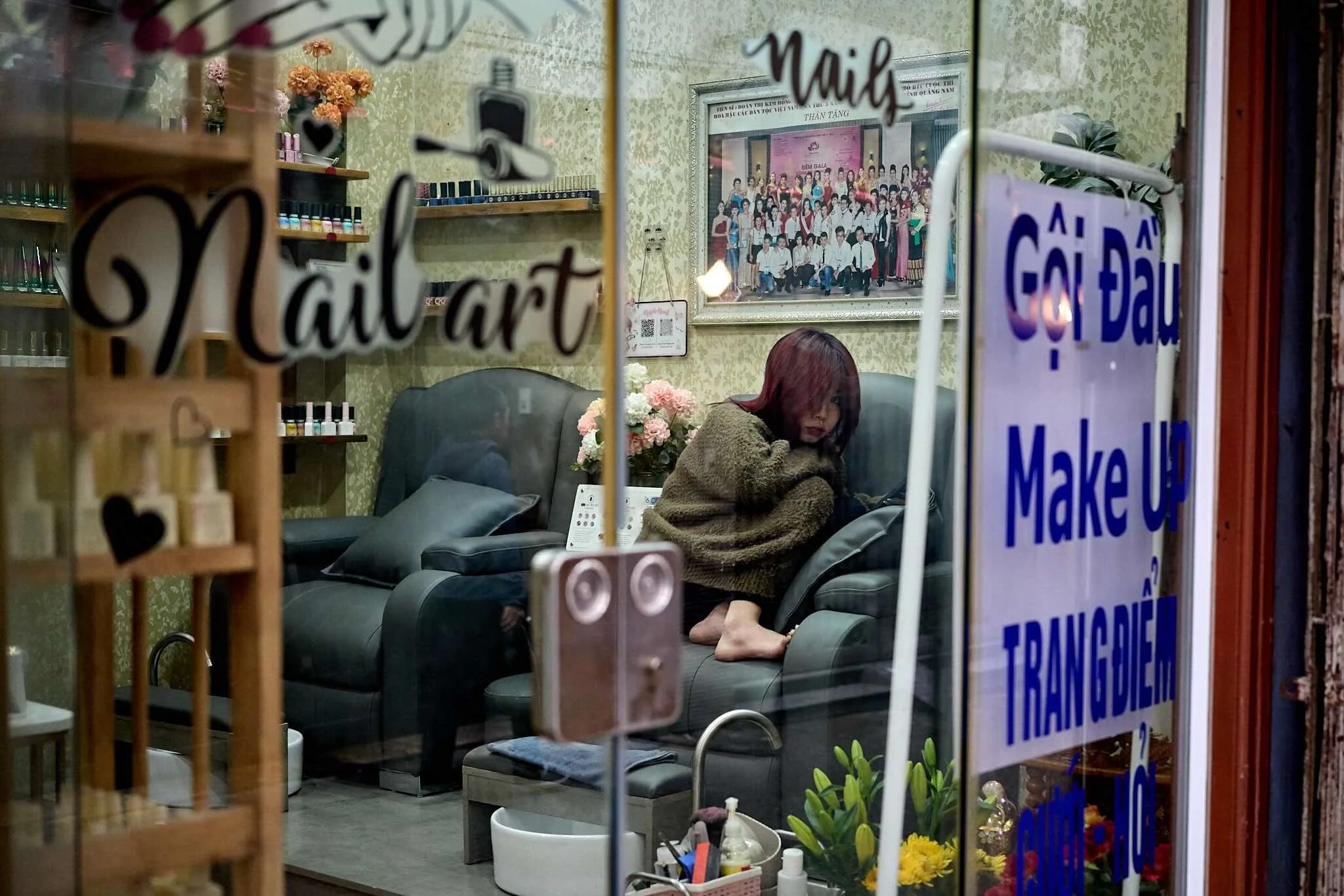 Reflection through a glass door shows a woman with reddish-brown hair sitting barefoot on a black leather sofa, with her legs curled under her, in a cozy room decorated with flowers, framed photographs, and floral wallpaper. A large group photo is vi