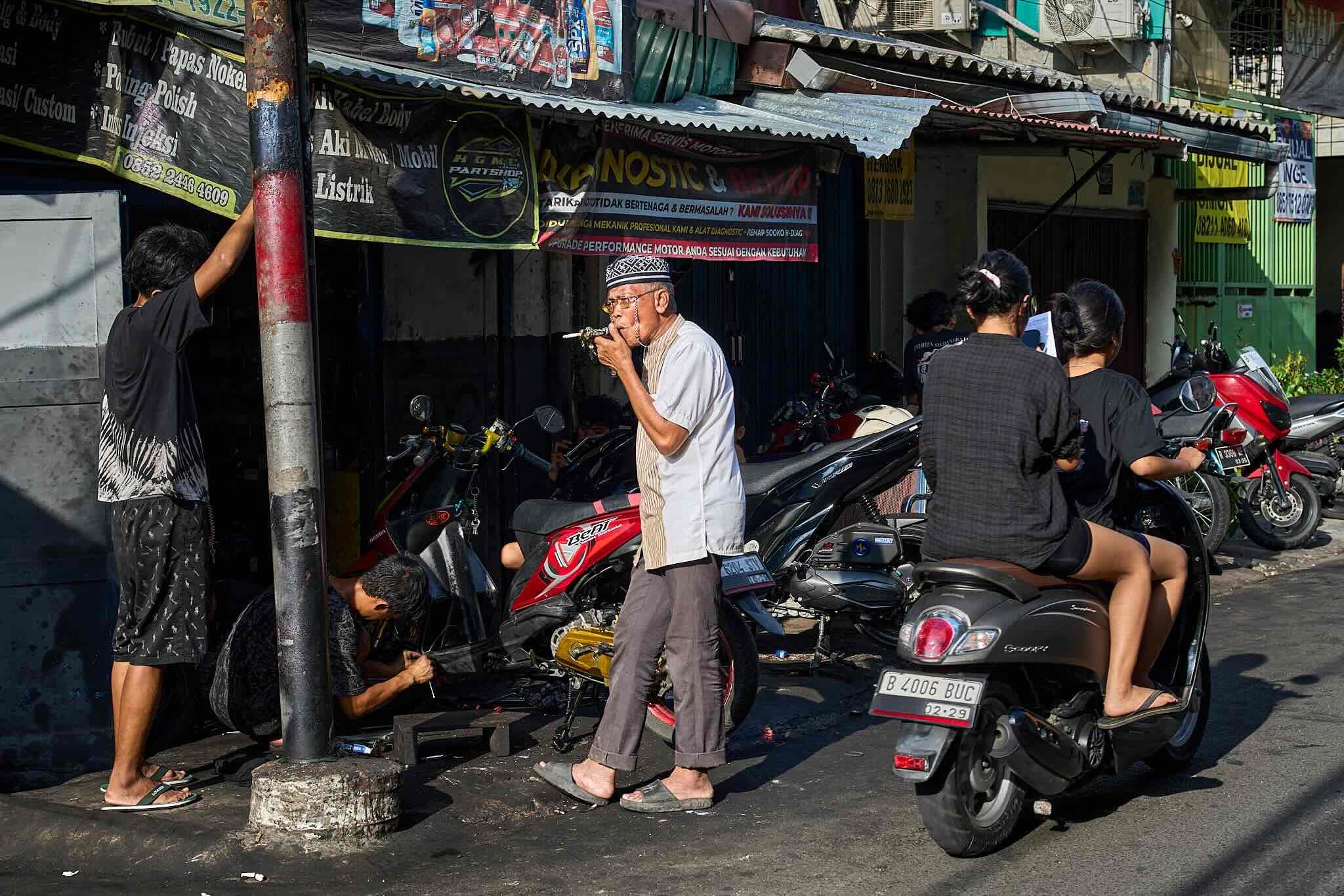 A busy street scene with several motorbikes parked, and multiple people gathered around. An older man wearing a traditional cap is playing a wind instrument, surrounded by women and men, some on motorcycles and others standing. Buildings with signs a
