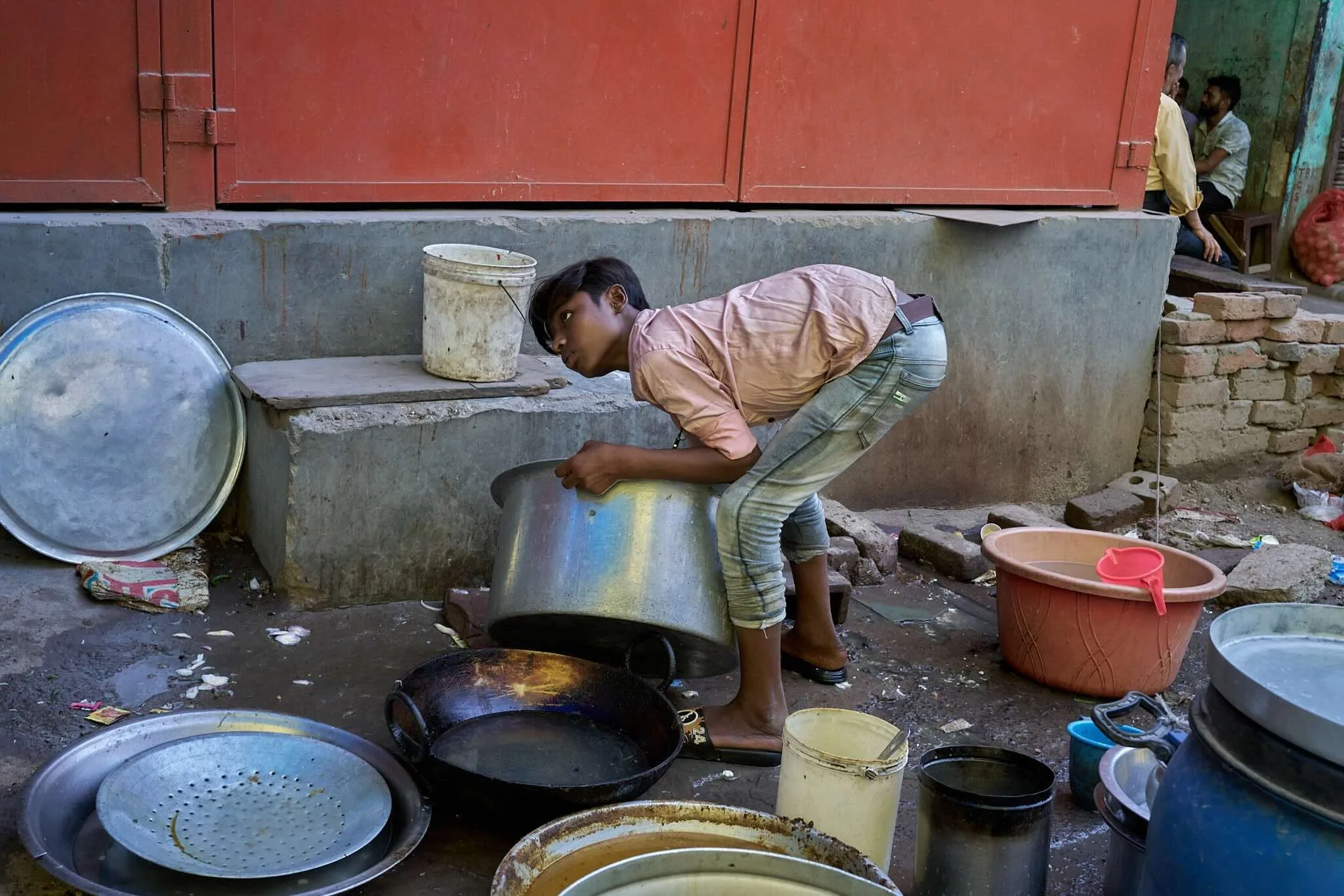 Jerzy Jaworek, Street and Travel Photography, India, Delhi. A boy is washing dishes outdoors in a makeshift area with buckets, pans, and utensils scattered around. Other children sit in the background near a brick wall and green-painted wall.