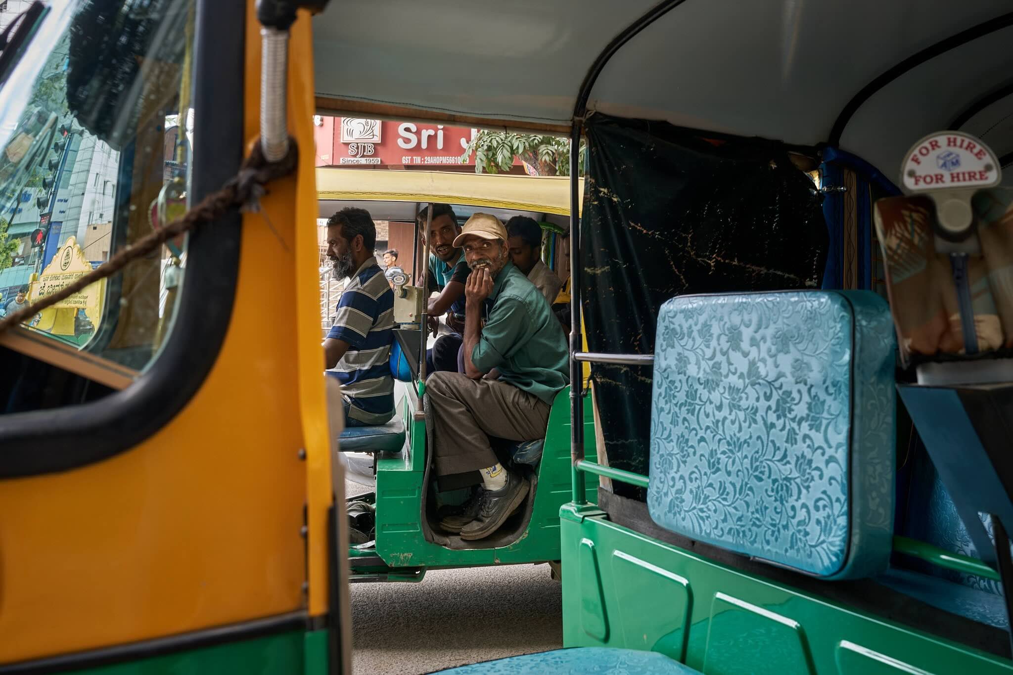 Jerzy Jaworek, Street and Travel Photography, India, Delhi. View inside a colorful auto rickshaw with several men seated, looking towards the camera, with signs and street scene visible outside.