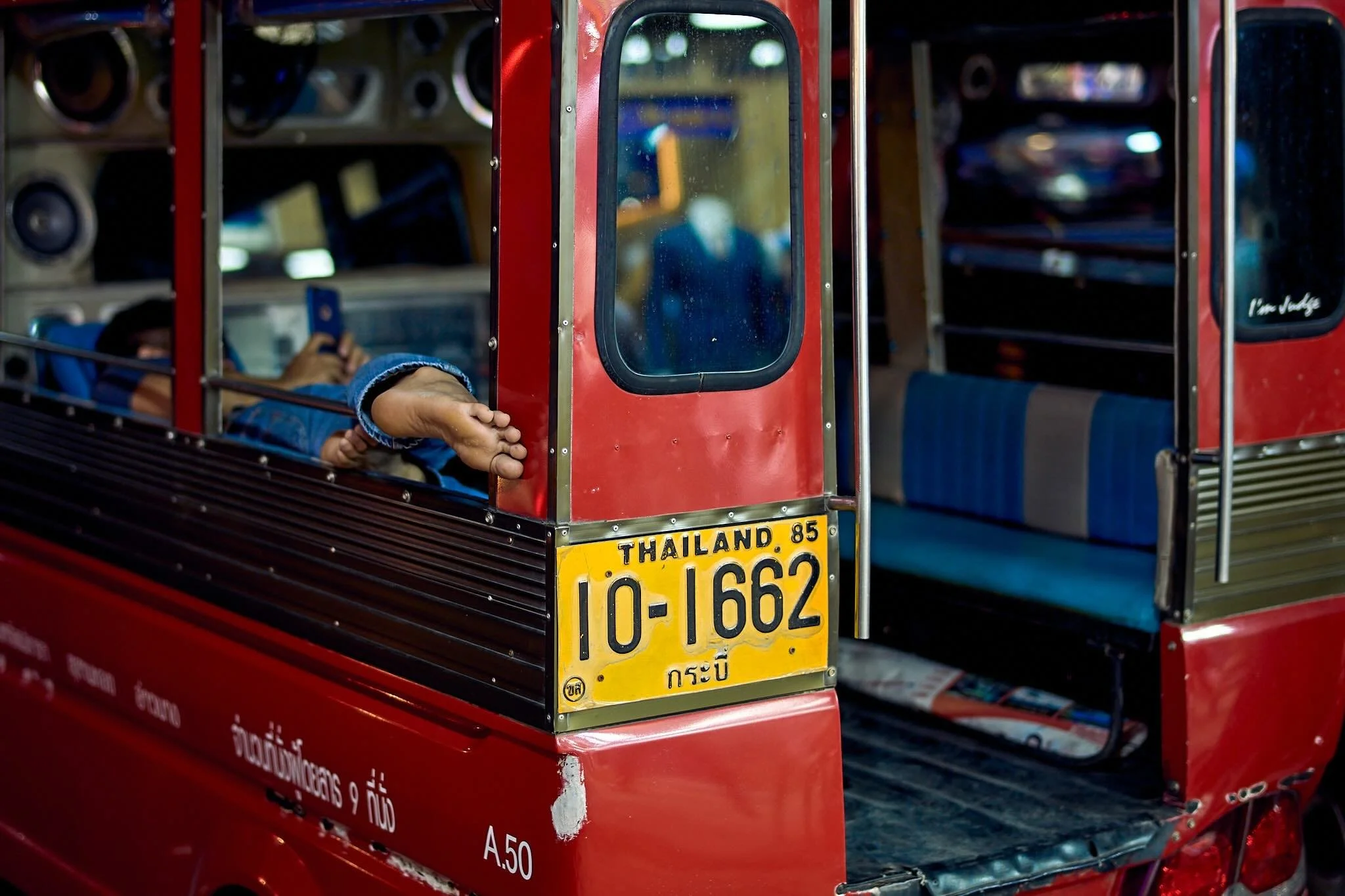 A red tuk-tuk parked, with a passenger sitting inside using a smartphone, Thailand license plate, and Thai characters on the side.