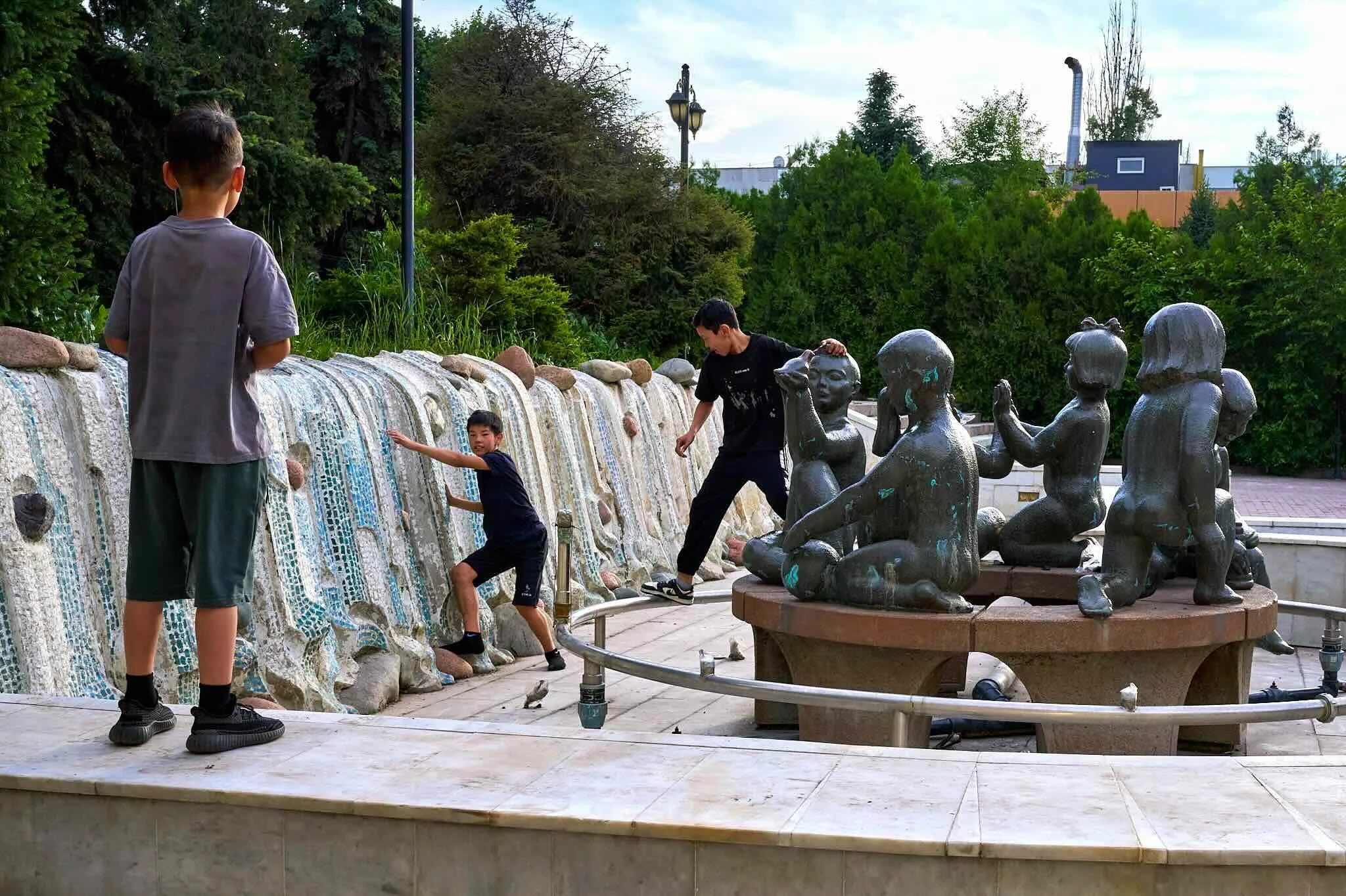 Jerzy Jaworek, Street and Travel Photography, Kazakhstan, Almaty. Four boys playing near a water feature with bronze statues, a fountain, trees, and a cloudy sky in the background.