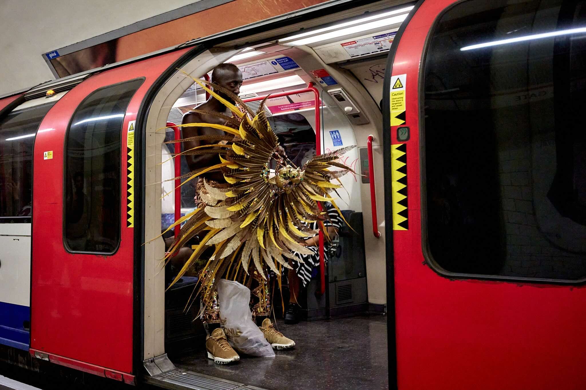 A person dressed in an elaborate gold and black costume with feathers, sitting inside a red and black subway train.