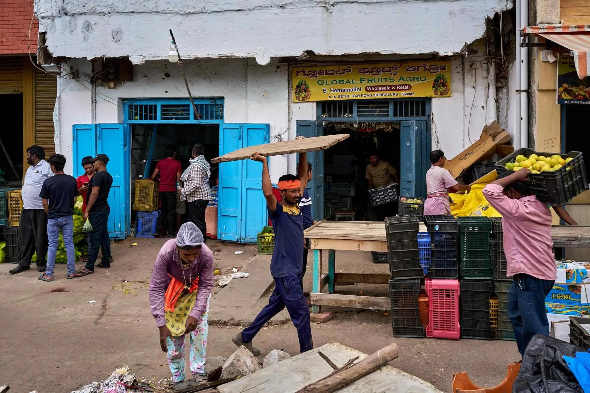 Jerzy Jaworek, Street and Travel Photography, India, Delhi. Street scene showing people working in a market with fruit stalls, crates, and baskets, in front of a store with a yellow signboard reading 'Global Fruits Agro' in Bengaluru.