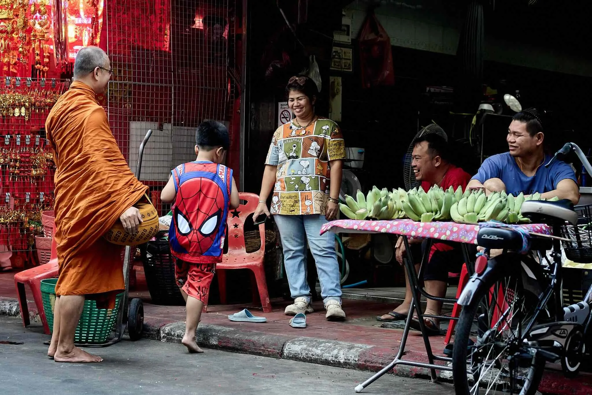 Group of people at street market, including a monk, a boy with Spider-Man backpack, a woman smiling, and two men sitting, with bananas on table, parked bicycles and market stall in background.